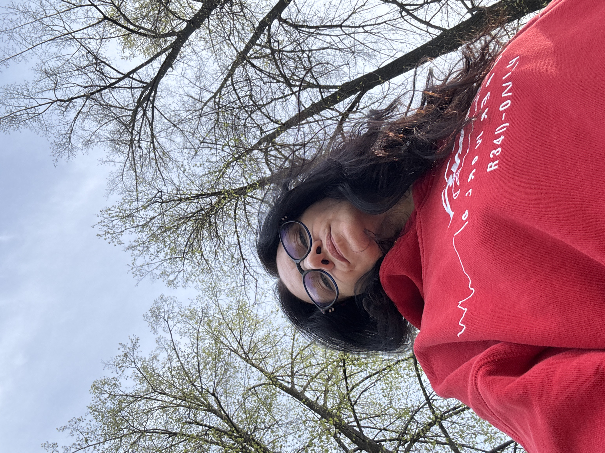 A woman with dark hair and glasses is pictured from a low angle, looking directly at the camera. She is wearing a red hooded sweatshirt with white writing on the sleeve. Behind her, tree branches with small green buds are visible against a light, overcast sky. The location appears to be outdoors, possibly a park or wooded area. Without additional context, it is difficult to confirm the location in Chaikovsky, Russia.