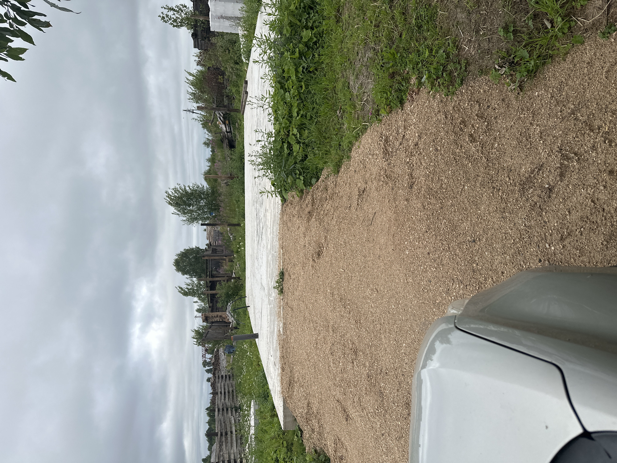 The image shows an outdoor scene in Plesetsk, Russia, with a focus on a newly constructed concrete foundation. The foundation is a light gray, rectangular slab, elevated slightly above the surrounding ground. A vertical gray pipe protrudes from the center of the foundation.
In the foreground, a path of light brown gravel leads towards the foundation. The edge of a silver car is visible in the lower left corner of the frame, partially obscuring the gravel path and suggesting the image was taken from within or near the vehicle.
The surrounding environment is characterized by greenery and a cloudy sky. Lush green grass and various plants, including some with broad leaves, grow around the foundation and along the edge of the gravel path. In the background, there are wooden fences, stacks of lumber, and some trees and bushes, indicating a rural or semi-rural setting. A faint structure, possibly part of a bridge or industrial equipment, is visible in the distance. The sky is overcast with gray clouds, suggesting a mild or cool day.