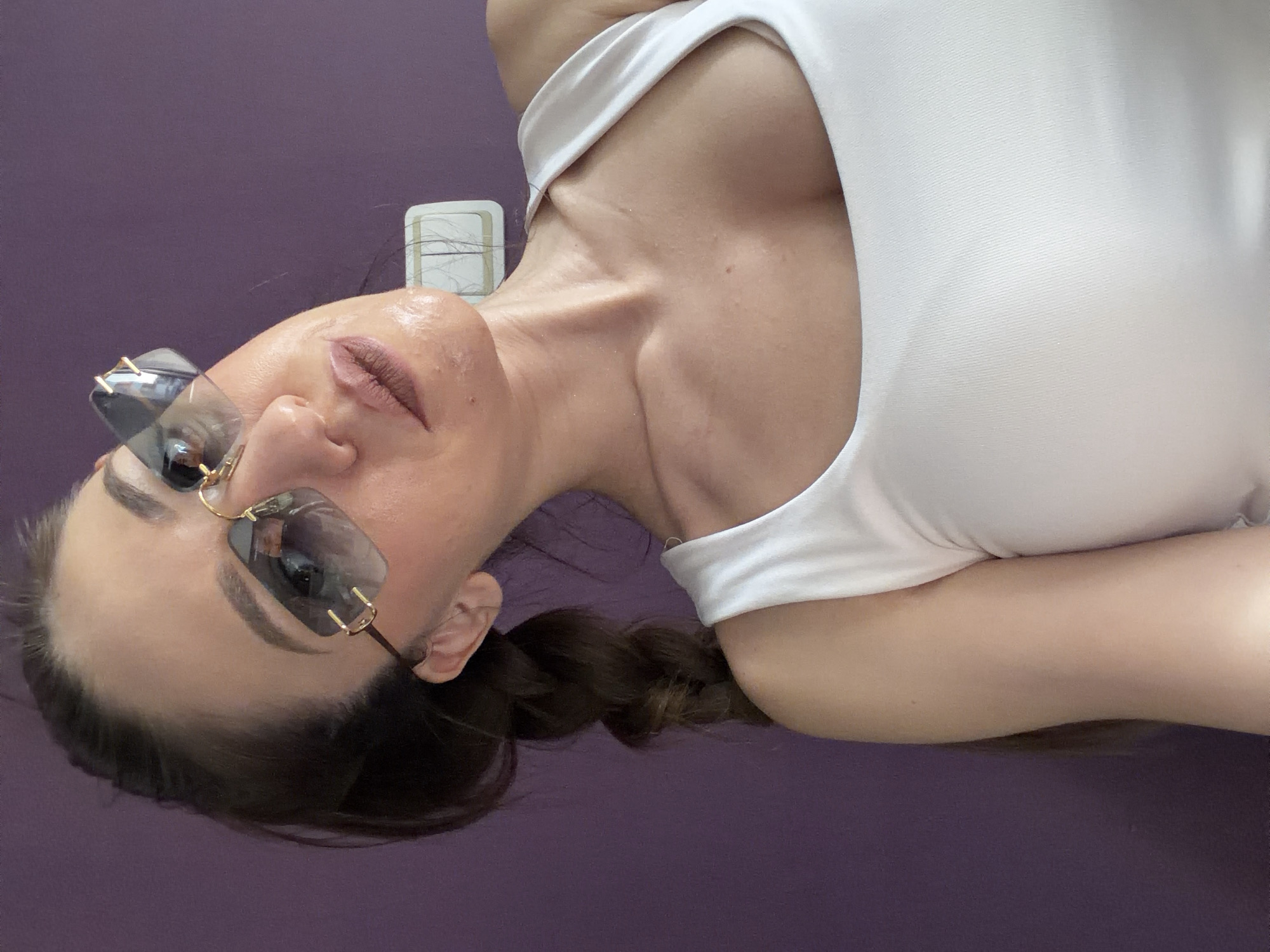 The image displays a close-up, chest-up portrait of an adult female subject, positioned at an upward angle towards the camera. Her dark hair is styled in a braid that extends over her left shoulder. She is wearing rimless sunglasses with gradient gray lenses and gold-colored metallic frame details, obscuring her eyes. Her lips are accentuated with a matte, neutral-toned lipstick, and her facial skin, particularly around the forehead, nose, and upper lip, exhibits a visible sheen. The subject is dressed in a white, form-fitting scoop-neck tank top. The background consists of a uniformly colored purple wall. On the wall, to the right of the subject's head, a white rectangular electrical faceplate housing two vertical switch mechanisms is visible. This electrical fixture is consistent with standard installations found in European urban environments, including the city of Sofia, Bulgaria, where the scene is located. The subject's posture indicates she is posing for a photograph.