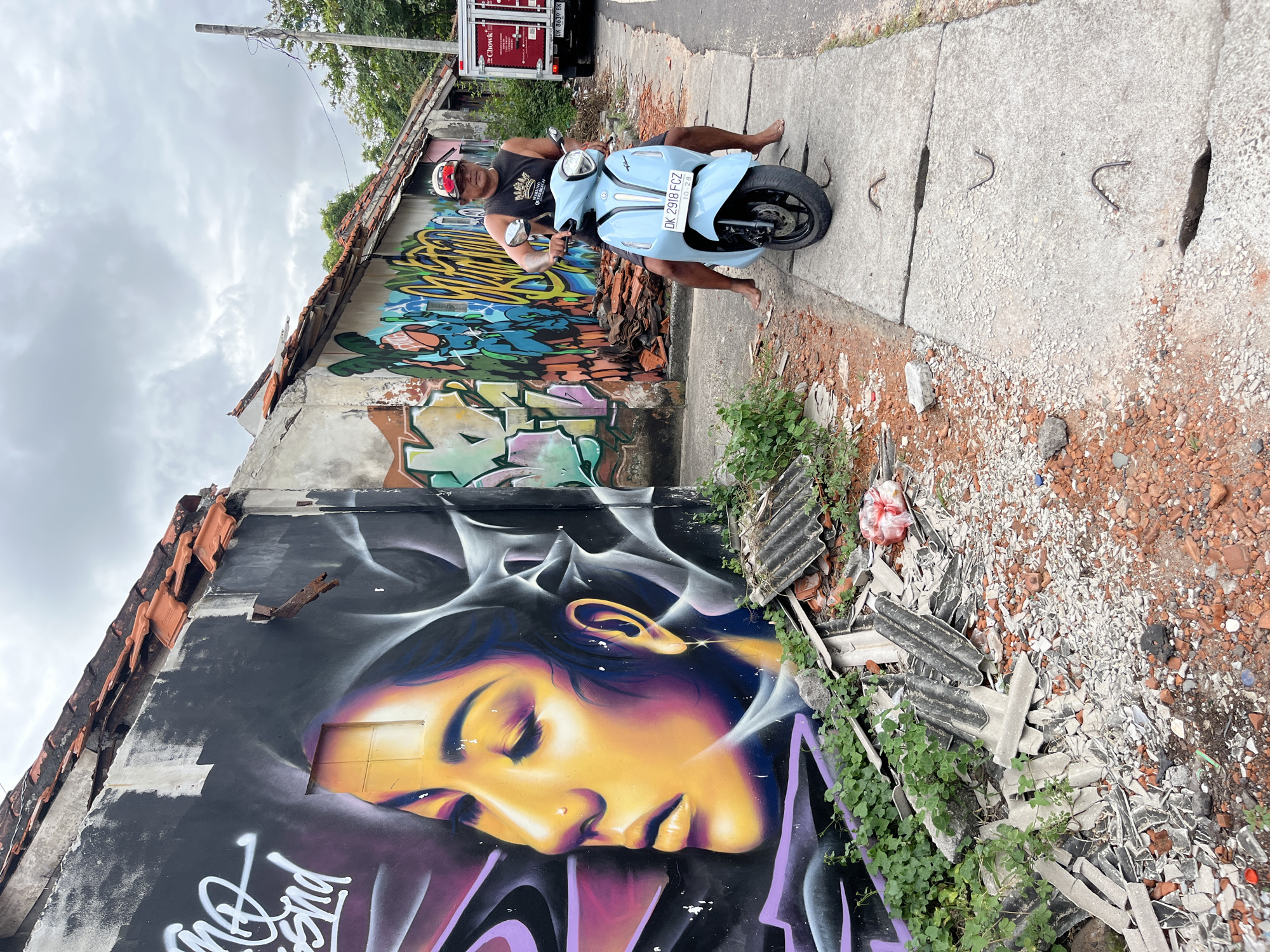 A man wearing a black tank top and a red baseball cap is sitting on a light blue scooter on a concrete sidewalk in Kuta Selatan, Indonesia. He is facing the camera and has his feet on the ground. The scooter has a license plate that reads "DK 2918 FCZ". Behind him, a wall is covered in graffiti, featuring a large mural of a woman's face with vibrant colors. The building appears to be in disrepair, with broken roof tiles and debris scattered on the ground. To the right of the man, there is a red truck with the word "Chowk" visible on its side. The sky is overcast with clouds.