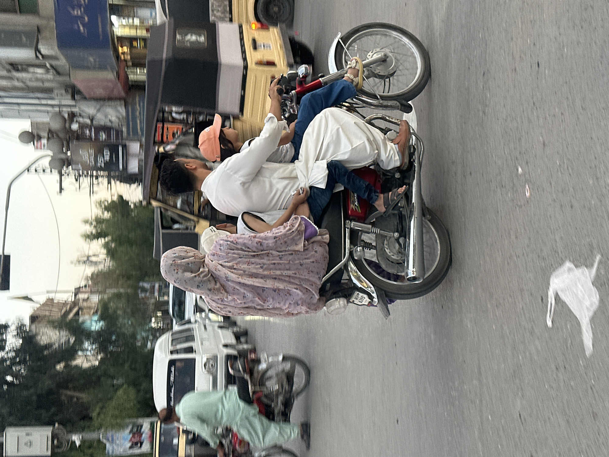 A scene unfolds on a street in Quetta City Tehsil, Pakistan, where a family of four navigates traffic on a motorcycle. A man, dressed in traditional white shalwar kameez, operates the motorcycle. Seated behind him is a young girl wearing a peach-colored baseball cap. Between the man and the girl, a woman is positioned, her head covered in a light-colored, patterned headscarf. On the very back of the motorcycle, another child, wearing a white shirt, is seated. The family is captured in motion, amidst other vehicles including a white van to their left and a yellow auto-rickshaw to their right, suggesting a typical urban street environment with moderate vehicle density. The road surface appears paved, and the surroundings include buildings and trees, indicating an urban setting. A piece of white plastic debris is visible on the road in the foreground. The lighting suggests it is daytime, possibly late afternoon or early evening.