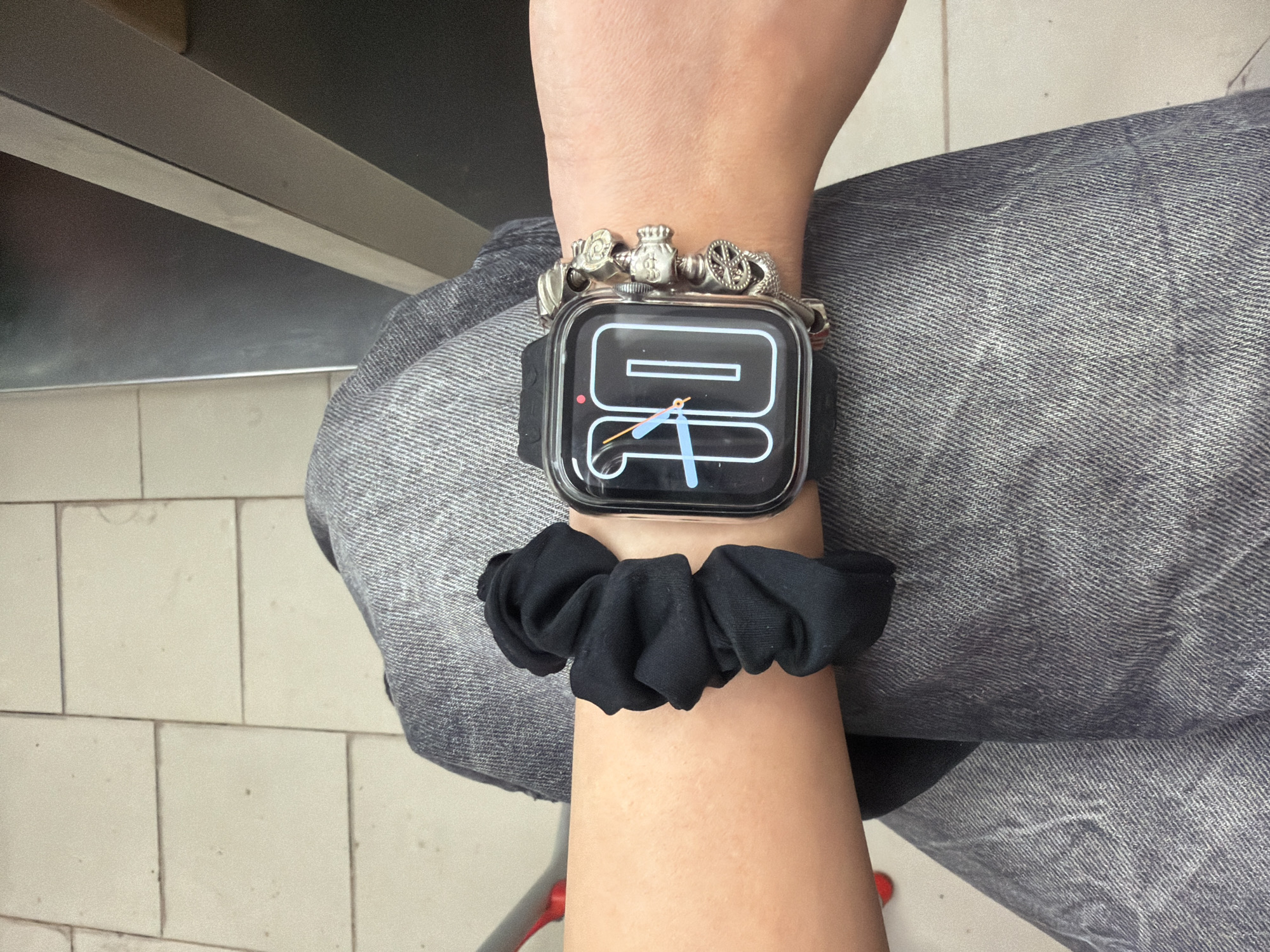 The image shows a close-up of a person's arms and legs, clad in denim jeans. On the left arm, a black scrunchie is visible. On the right wrist, a smartwatch displays the time "10" with a blue hour hand and an orange minute hand. A bracelet with multiple silver charms, including skull and circular designs, adorns the same wrist. The background consists of light-colored tiled flooring and a metallic surface. The scene does not provide any specific context that would allow for its location to be identified in the city of Plovdiv, Bulgaria.