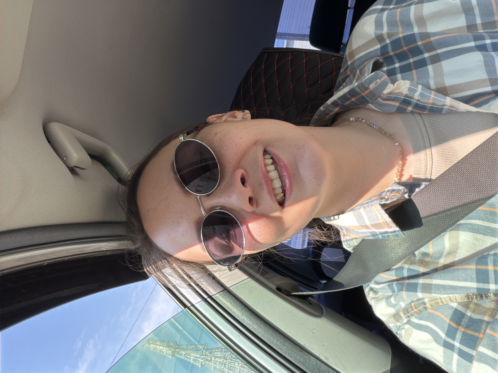 A young woman with brown hair, wearing sunglasses, a beige t-shirt, and a plaid shirt, is sitting in a car. She has freckles and is smiling. A seatbelt is across her chest. Outside the car window, a tall, lattice-like tower is visible against a blue sky with scattered clouds. The interior of the car includes a gray headliner, a beige grab handle, and a black seat with red stitching. The woman is wearing a necklace. The scene is illuminated by sunlight, casting shadows.