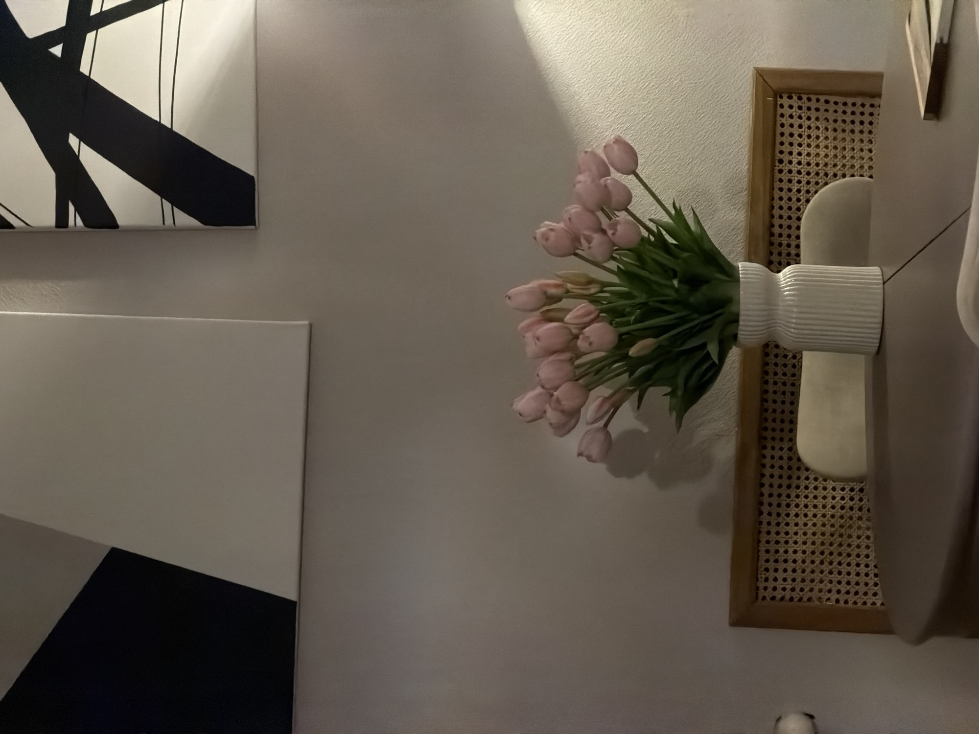 The image depicts a still life arrangement on a wooden dining table. A white, ribbed vase holds a bouquet of pink tulips, their stems and green leaves visible. Behind the vase, the rattan back of a chair is partially obscured by the table. Two abstract paintings hang on a textured, neutral-colored wall. The painting on the left features large geometric shapes in black and white, while the painting on the right is composed of intersecting black lines on a white background. The lighting suggests an indoor setting, possibly a dining room, with light casting a shadow from the tulips onto the wall. The image does not contain any discernible elements that would indicate a specific location in Moscow, Russia.