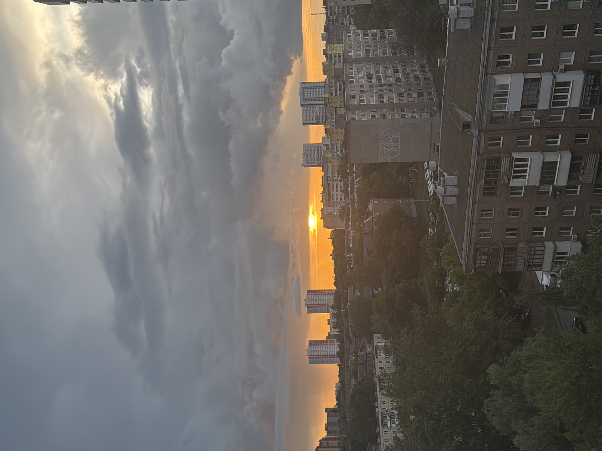The image displays a dramatic sunset over a densely populated urban landscape. The sky is a prominent feature, dominated by heavy, dark grey clouds on the left and upper portions, suggesting dynamic weather, possibly after or before a storm. On the right, a brilliant golden sun is visible low on the horizon, casting an intense orange and yellow glow across the lower sky and illuminating the distant clouds.
Below the horizon, an extensive cityscape unfolds, characterized by numerous multi-story residential buildings. These include older apartment blocks with repetitive window structures, some featuring enclosed balconies and visible air conditioning units. Further in the distance, taller, more modern-looking buildings with distinct square window patterns can be observed. The architecture suggests a large, established city.
Abundant green trees and dense foliage are interspersed throughout the urban environment, appearing in the foreground, between buildings, and lining what appear to be roads or pathways. Some parked cars are faintly visible on these routes. The time of day is clearly sunset, marked by the low sun and the warm, golden light that bathes parts of the scene, creating a striking contrast with the darker clouds. No discernible text is visible within the image.