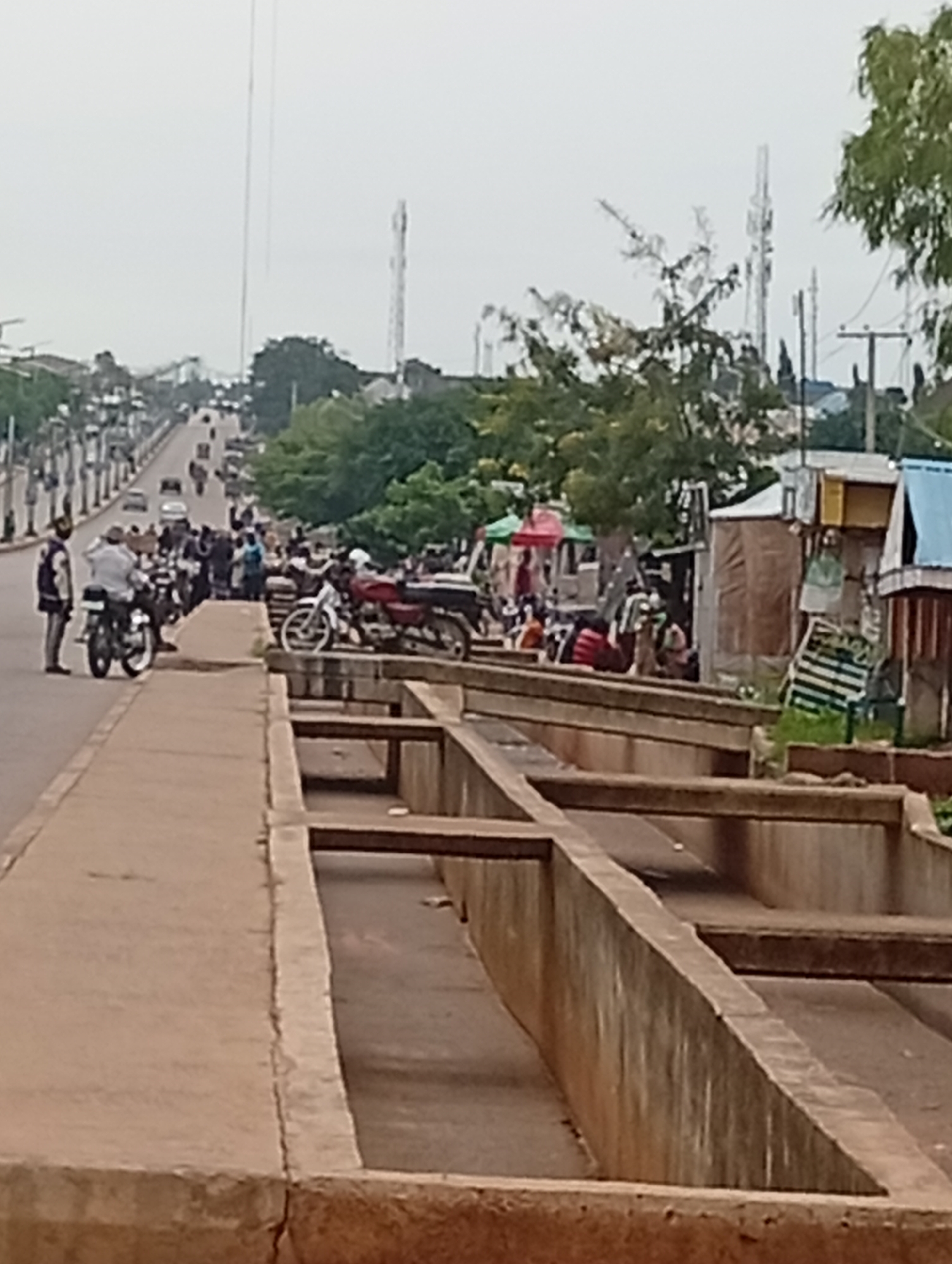 The image depicts a street scene in Tumfure, Nigeria. In the foreground, a concrete structure with multiple sections runs along the right side of the frame. To the left, a paved walkway is visible, leading to a road with several motorcycles parked along its edge. A few individuals are present, with one person standing next to a parked motorcycle. Further down the road, more motorcycles and vehicles are seen, along with a gathering of people. The background shows a street lined with trees and buildings, and in the distance, communication towers rise against an overcast sky. The overall atmosphere suggests a busy urban environment.