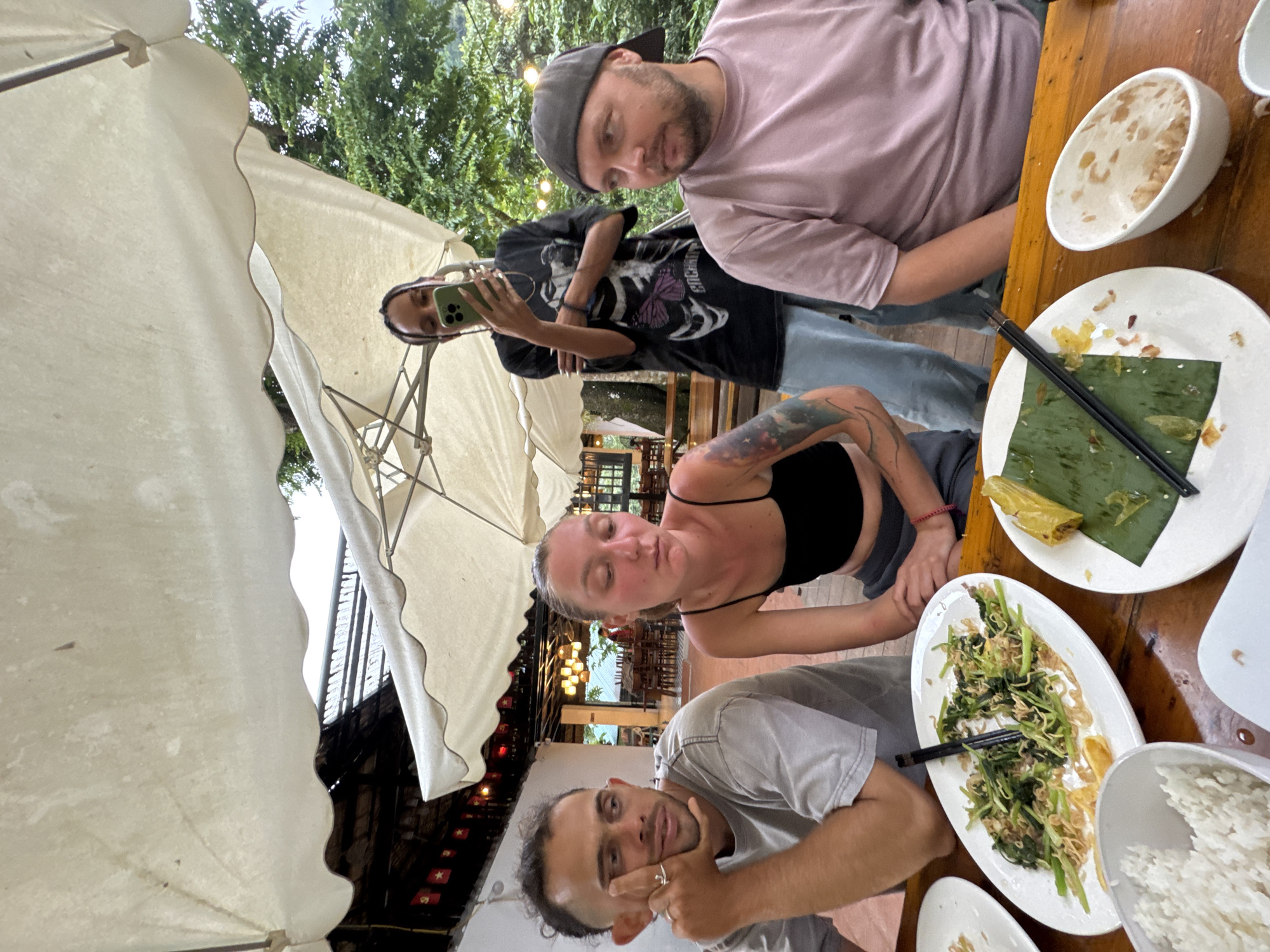Four people are seated at a wooden table under a large white umbrella. The background appears to be an outdoor restaurant in Cổ Lũng Commune, Vietnam, with trees and a building visible. In the foreground, there are plates of food, including rice and stir-fried vegetables. One person, a woman with dark hair in braids, stands and takes a selfie with her phone. The other three individuals are seated, appearing to be engaged in conversation or observing their surroundings. The lighting suggests it is daytime, possibly overcast or shaded.