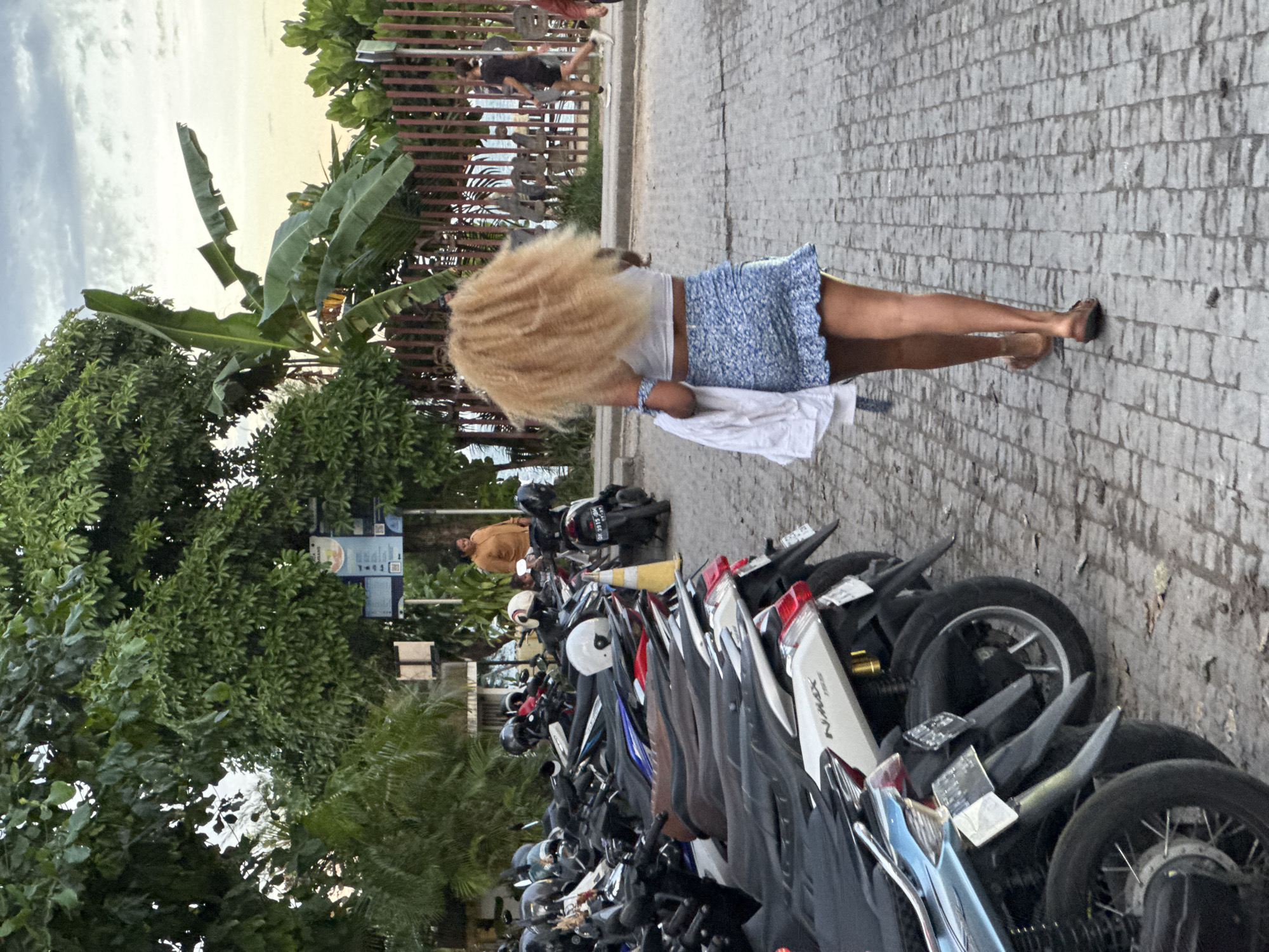 A woman with long, light-colored hair walks on a paved area beside a dense parking lot of scooters. She is wearing a white top and a blue floral skirt and carries a white cloth in her hand. The pavement is light grey, and there are trees and a fence to the left. In the background, people walk along a path near water. The scene is located in Denpasar, Indonesia, and includes vegetation typical of a tropical environment.