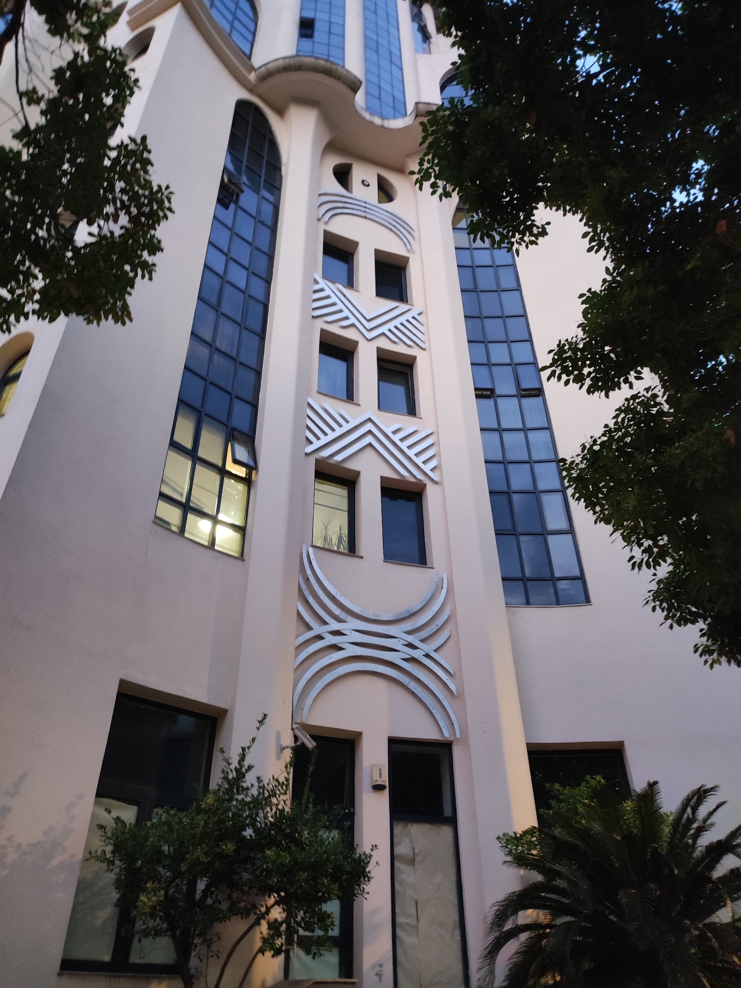 The image shows the exterior of a modern building with a beige facade and large, dark-tinted windows. The building has a distinctive architectural style with curved elements and decorative metallic accents. Trees with dark green foliage frame the building on the left and right sides, partially obscuring the upper floors. Some of the windows are open, and the light from inside is visible in one of them. The sky is visible through the trees and appears to be twilight or early evening. The building's design suggests it could be a commercial or residential building. Based on the architectural style and the presence of palm trees and lush vegetation, this building is likely located in Batumi, Georgia, a coastal city known for its contemporary architecture and subtropical climate. The image is taken from a low angle, looking upwards at the building. There are no people visible in the image.