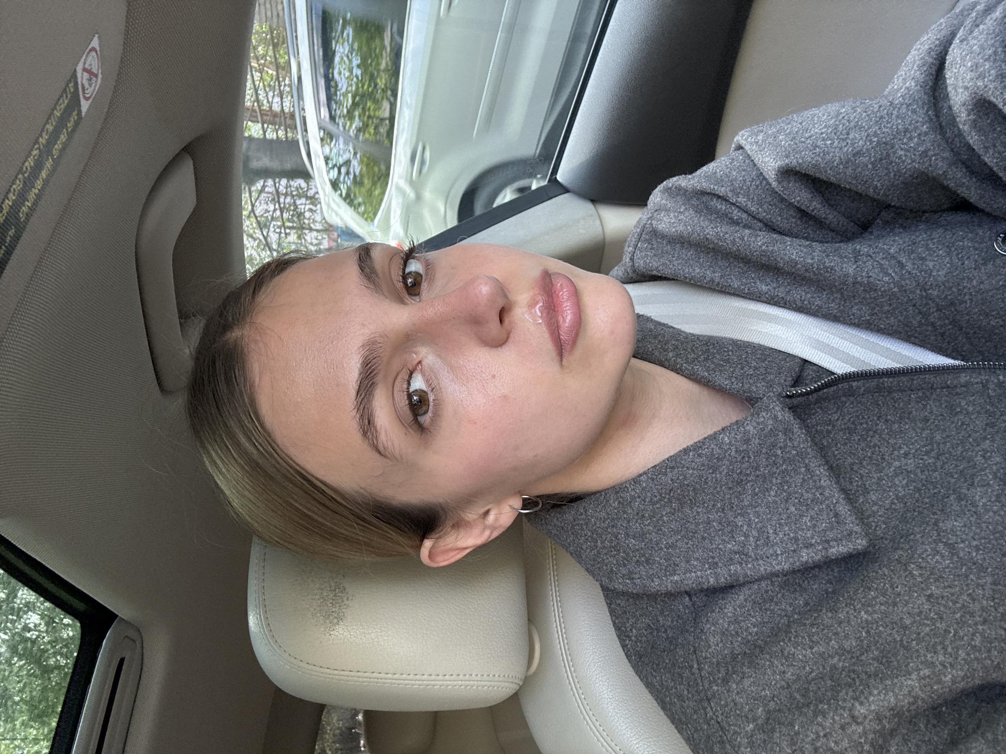 A medium shot depicts a light-skinned woman in a car. She has light brown hair and brown eyes, wearing a grey jacket with a white seatbelt across her chest. She has a neutral expression. The car interior is visible, including a beige headrest and the car window, which reflects a street scene with greenery and a white car. Based on visual cues like architecture and vehicle types, the location might be Tbilisi, Georgia. No specific landmarks or identifiers confirm this precisely, but the overall impression suggests an urban setting in that geographic region.
