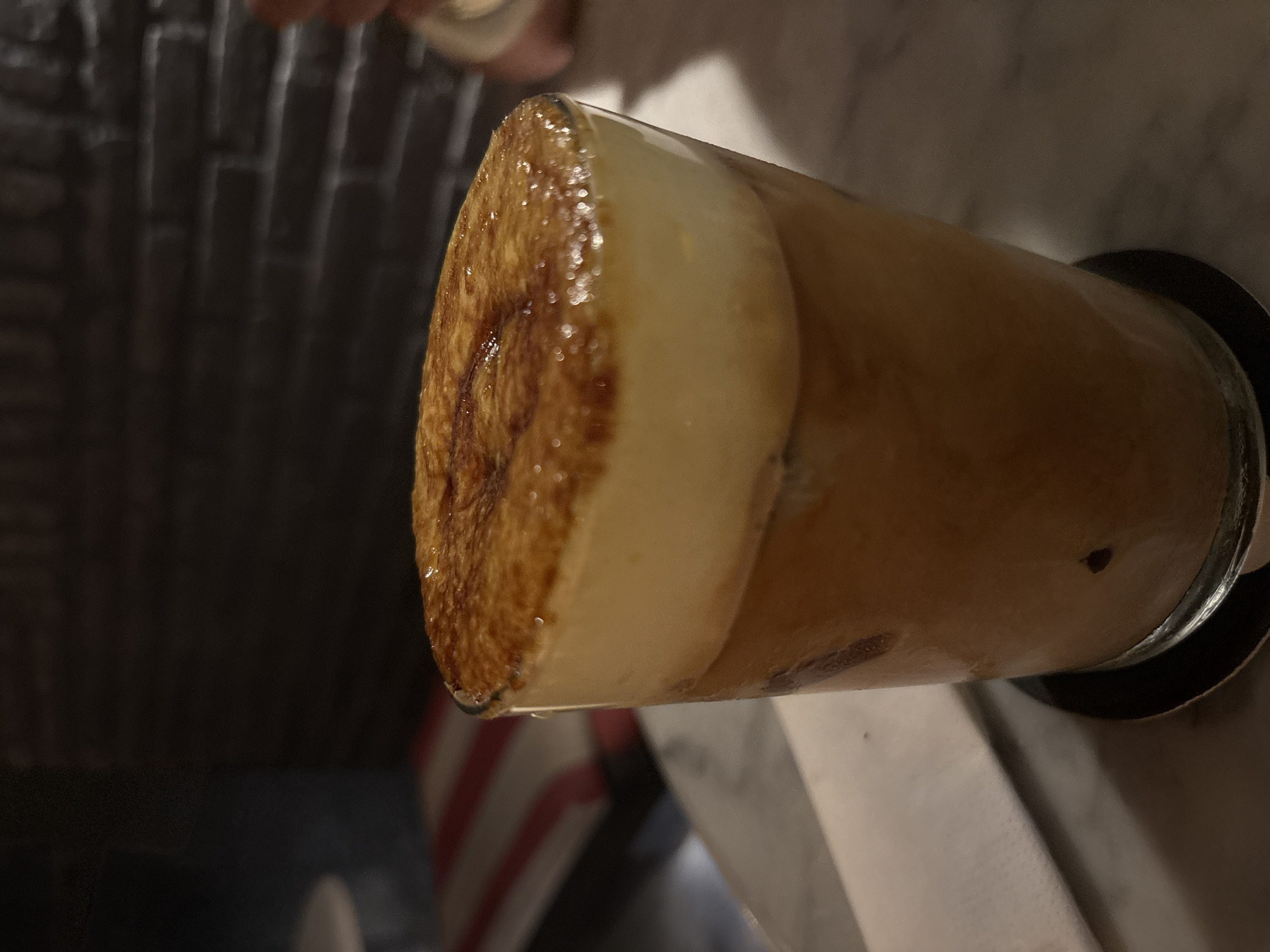 An iced beverage with a layer of cream on top and a caramelized sugar crust is presented in a clear glass on a marble table. The drink has a light brown color and contains ice cubes. A white napkin is placed to the left of the glass. In the background, a dark brick wall is visible, and to the left, a blurred area with red and white stripes can be seen. The scene is dimly lit, suggesting an indoor setting, possibly a cafe or restaurant in North Kuta, Indonesia.