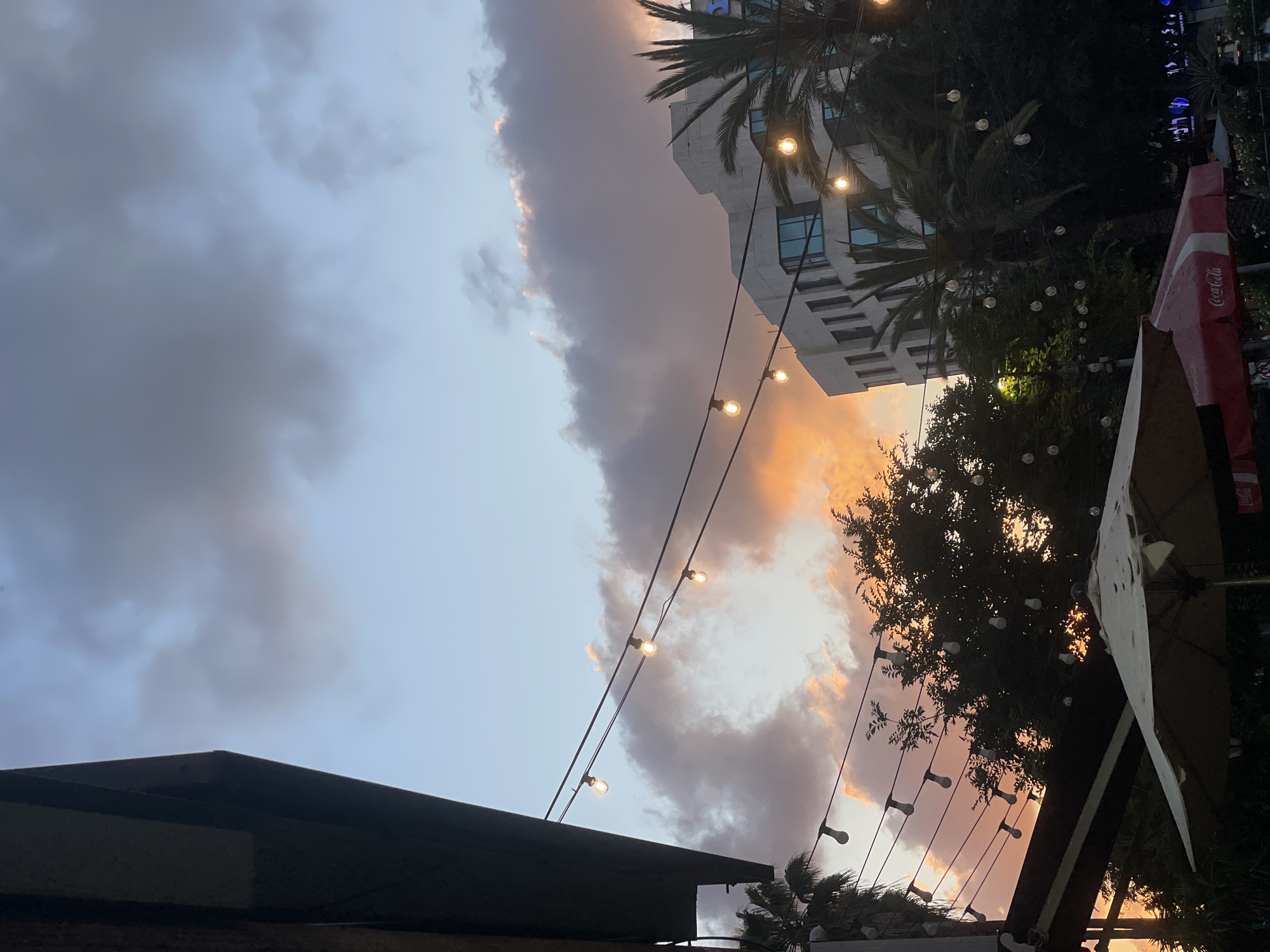 Captured at an outdoor setting in Raanana, Israel, the image shows a twilight scene, likely at a café or restaurant. An awning is visible in the lower part of the frame. Overhead string lights illuminate the setting. Palm trees frame the image, with a modern building in the middle ground. A Coca-Cola branded umbrella is on the right side of the frame. The sky, filled with dramatic clouds, shows hues of orange indicating sunset. No people are directly visible.
