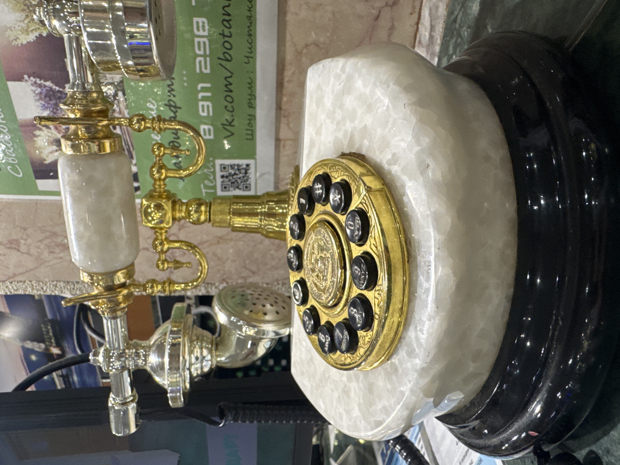 The image shows a close-up of an ornate, vintage-style rotary dial telephone. The telephone is predominantly gold-plated and features white marble accents on its base and handset. The rotary dial is also gold and has black buttons, with numbers and letters visible on some of them, including "0 OPER" and "#". The handset is supported by a gold cradle. In the background, a green paper with text and a QR code is visible, suggesting a commercial setting. Some of the visible text in Russian includes " vk.com/botanic" and "Шоу рум: Чистак". The overall impression is of a decorative item displayed in a shop or exhibition, possibly in Saint Petersburg, Russia, given the location context. The lighting is artificial and appears to be from overhead. There are no people visible in the image. The focus is entirely on the details of the telephone and its immediate surroundings.