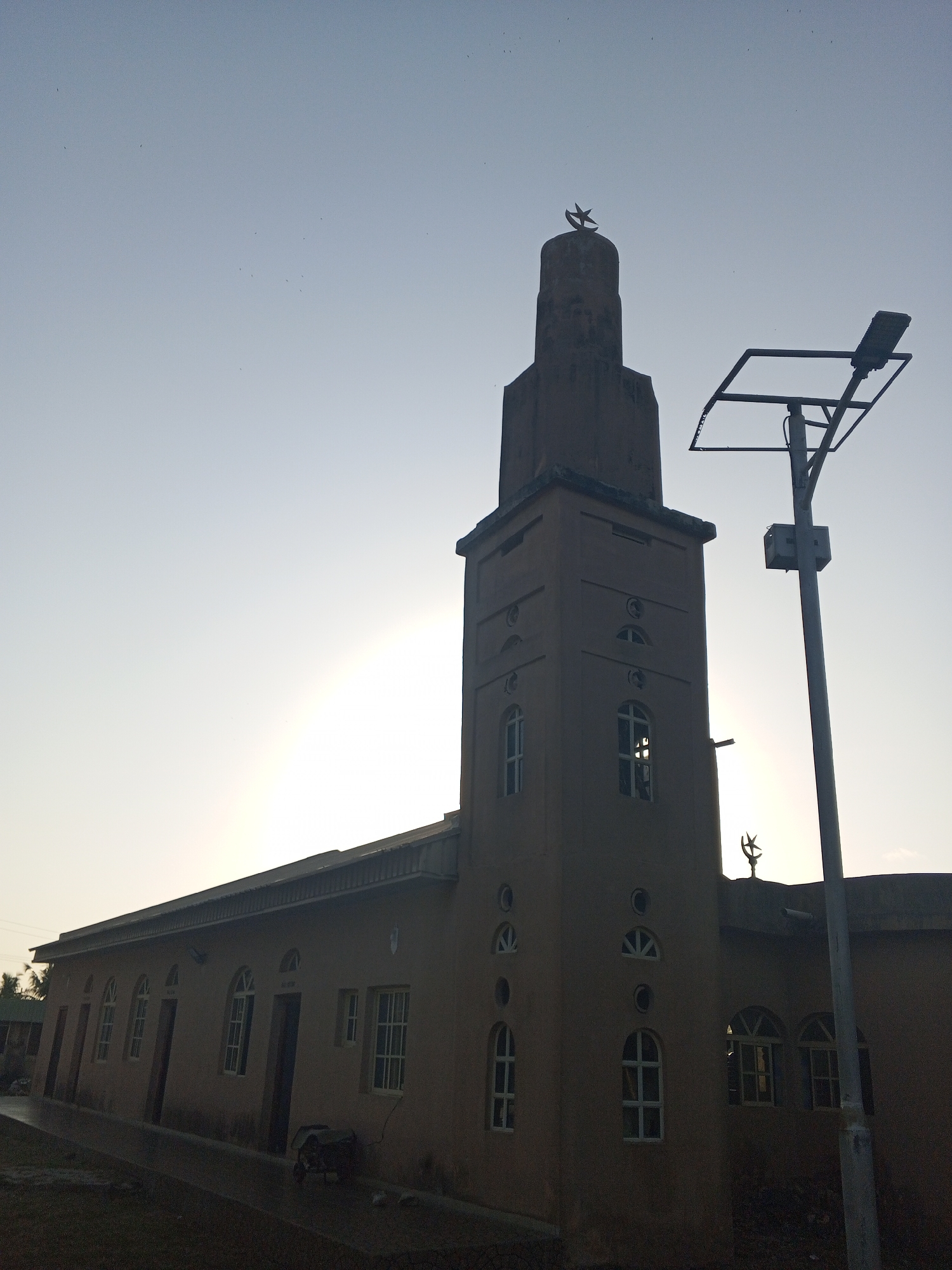 The image shows a mosque in Tawpo, Nigeria. The mosque is a single-story structure with a tall minaret. The minaret has a crescent moon and star on top. A street light is visible next to the mosque. The sky is light blue, and the sun is behind the building, creating a silhouette. The scene is quiet, with no visible people or activity.