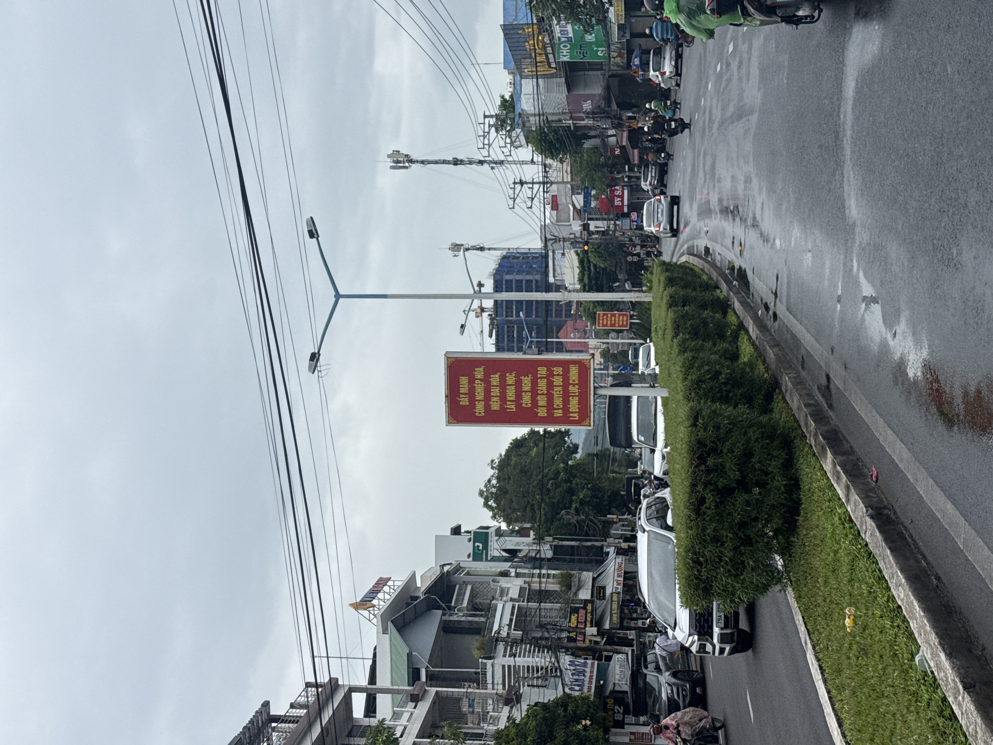 The image depicts a street scene in Nha Trang, Vietnam, on a cloudy day. The street is wet, suggesting recent rain. Traffic, including cars and motorcycles, is present, moving in both directions. Buildings line the left side of the street, displaying various signs and architectural styles. A prominent red sign with Vietnamese text is visible on the right side of the road. Green hedges and grass separate the lanes of traffic. Utility poles and overhead wires are also noticeable. In the foreground, on the grassy median, some yellow objects resembling toys are scattered.