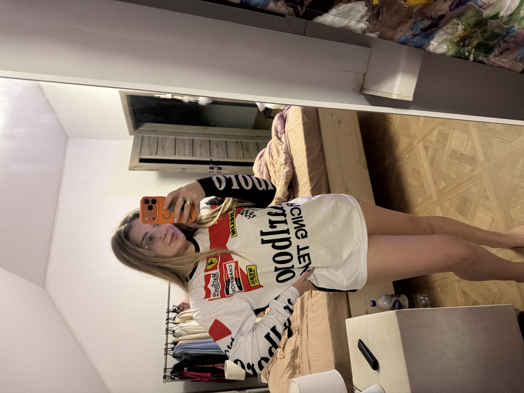 A young woman is captured in a mirror selfie, with the image rotated 90 degrees clockwise. She stands indoors, likely in a bedroom or dressing area located in Большая Шильна, Russia. She has long, light brown hair and wears glasses, looking towards the camera with a neutral to slightly smiling expression. She is holding an orange-cased smartphone with her right hand to take the photograph.
She is wearing a white long-sleeved shirt designed like a racing jersey, featuring prominent red and black sections. Visible text and logos on the shirt, which appear inverted due to the mirror reflection, include "FERRARI," "CHAMPION," "Marlboro" (partially visible), "RACING LEN," "POLO," and "MOTUL." Additional black lettering is present on the sleeves.
The background, reflected in the mirror, shows parts of the room. To the woman's right, a bed with a light brown headboard and a rumpled pinkish-purple blanket is visible, along with another patterned blanket. The floor is made of light wooden parquet. A light-colored, cube-shaped bedside table or dresser is positioned on the right side of the mirror, holding a dark object, a small water bottle, and a glass. To her left, a clothing rack filled with neatly folded clothes can be seen. The walls and ceiling are white. A small, open wooden structure, possibly a window or decorative vent, is reflected in the upper right corner of the mirror. The indoor lighting is diffused, providing a soft illumination to the scene.