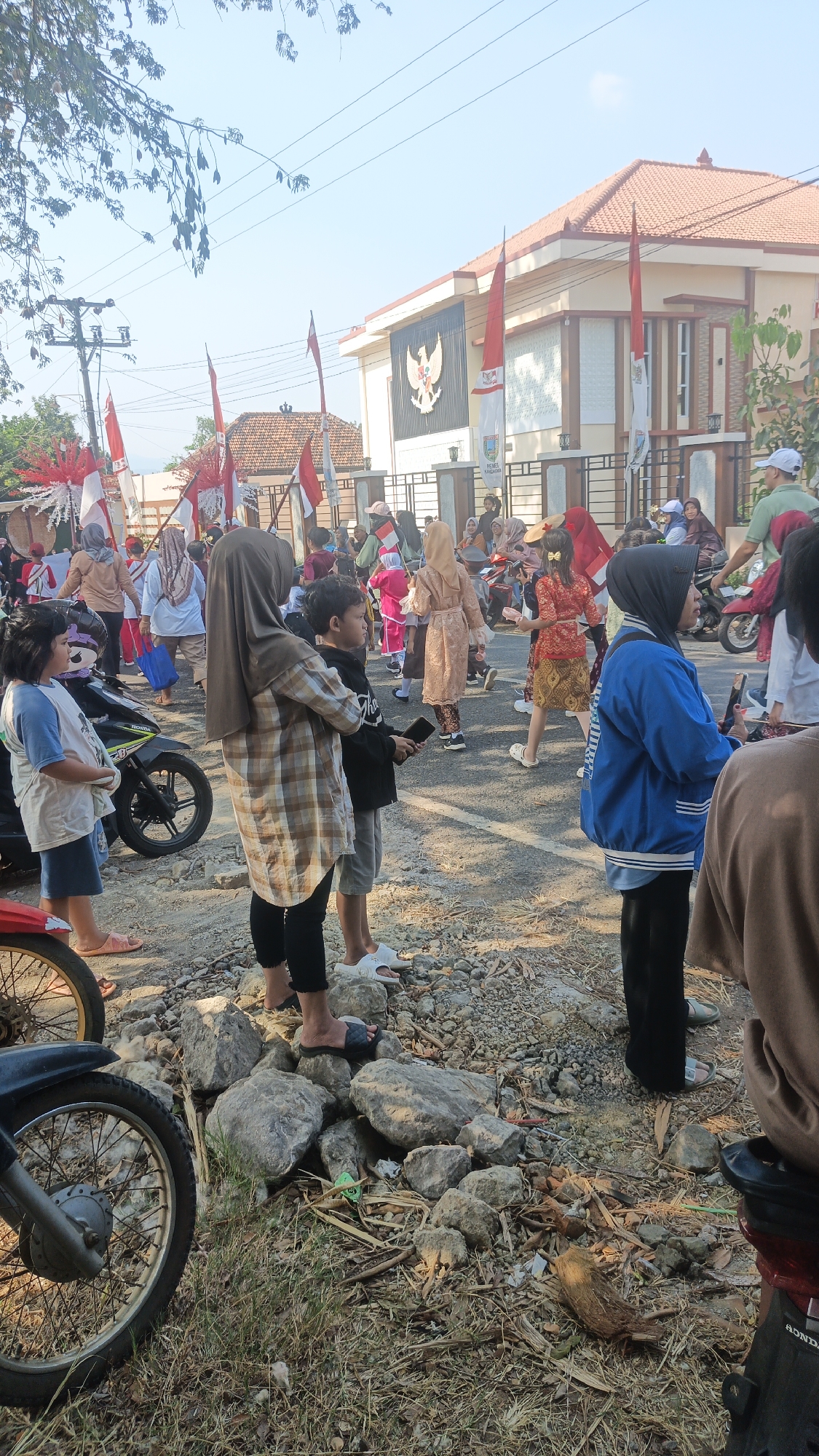 In an outdoor, daytime scene, a group of people, including children and adults, are gathered on a street. The street is paved and has a dirt and rocky shoulder. A building with a red-tiled roof and white walls is visible in the background, adorned with Indonesian flags. People are dressed in casual clothing, with some wearing headscarves. A motorcycle is parked on the left side of the image, and another is partially visible on the right. Some individuals are holding flags and other festive decorations. The atmosphere appears to be that of a public gathering or celebration.
