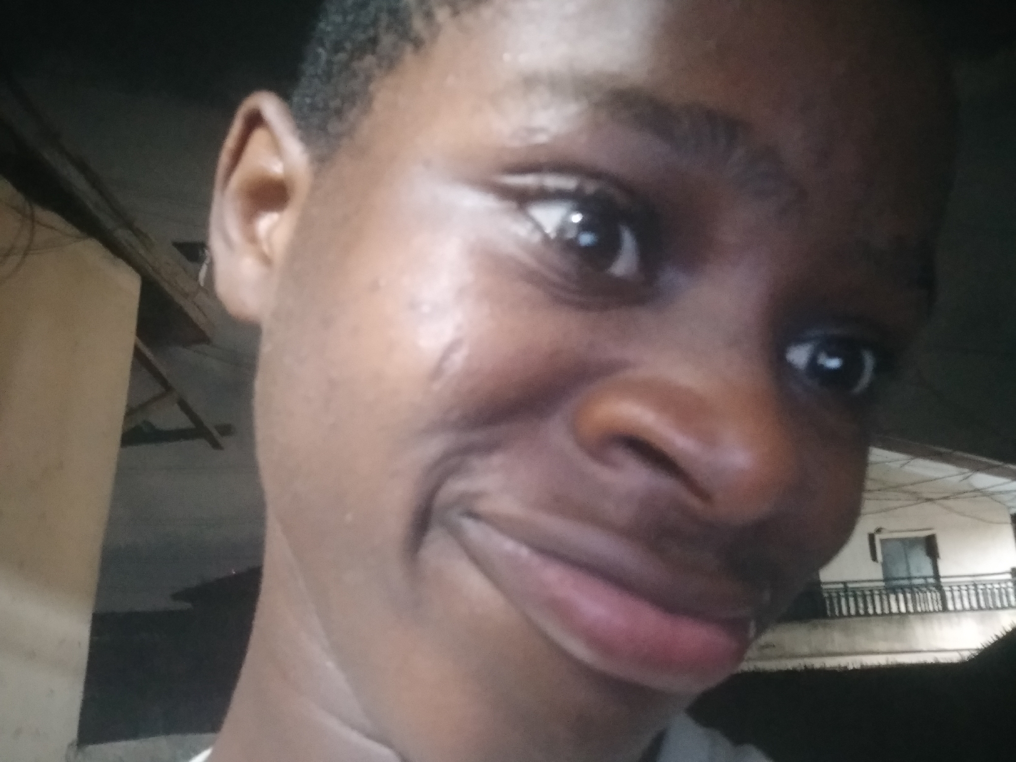 A close-up selfie of a dark-skinned individual with short, dark hair. The person is looking slightly to the right with a slight smile. Visible in the background is a building with a balcony, presumably in an urban environment. The location is Shangisha, Nigeria.