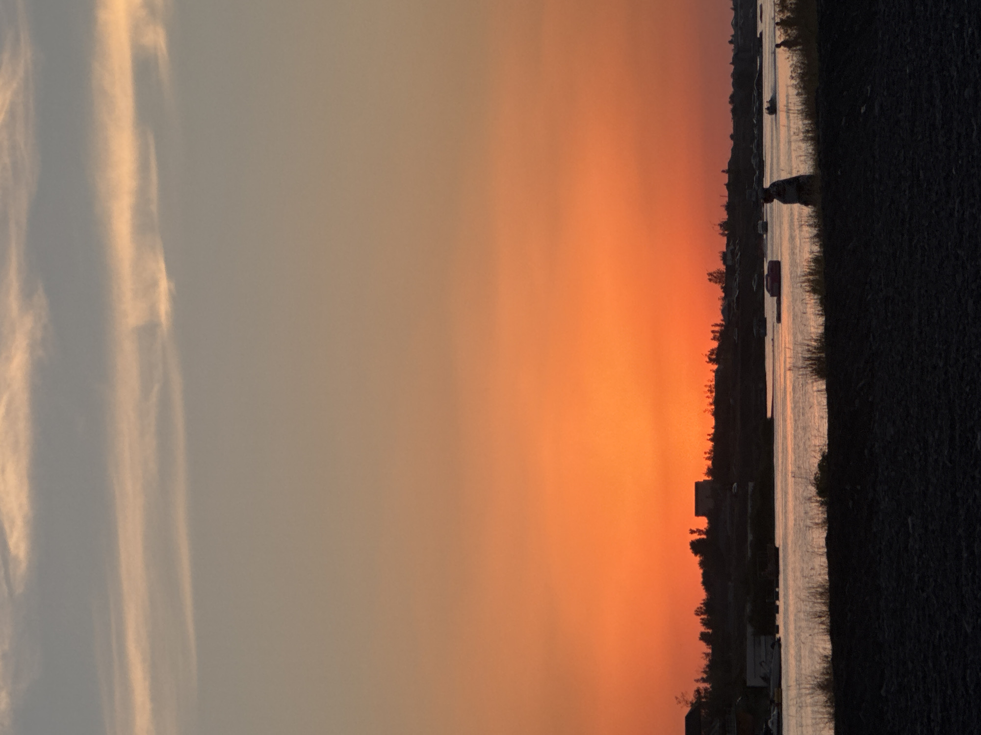 The image depicts a sunset over a body of water, likely in or near the village of Ақши, Kazakhstan. The sky is a vibrant gradient of orange and pink hues, with soft, wispy clouds stretching across the upper portion. In the foreground, a dark, gravelly embankment runs along the bottom of the frame. Beyond this, a calm body of water reflects the colors of the sky.
On the far bank of the water, a silhouetted treeline is visible, dotted with a few structures, including what appears to be a building. Scattered across the water are several small boats or buoys.
In the middle ground, near the shore on the right side of the image, a lone figure stands in silhouette, facing away from the camera and towards the water. Their posture suggests they are observing the sunset or the water. Further to the right, another smaller silhouetted figure is visible, closer to the water's edge. The overall scene is peaceful and serene, characteristic of a quiet evening by the water.