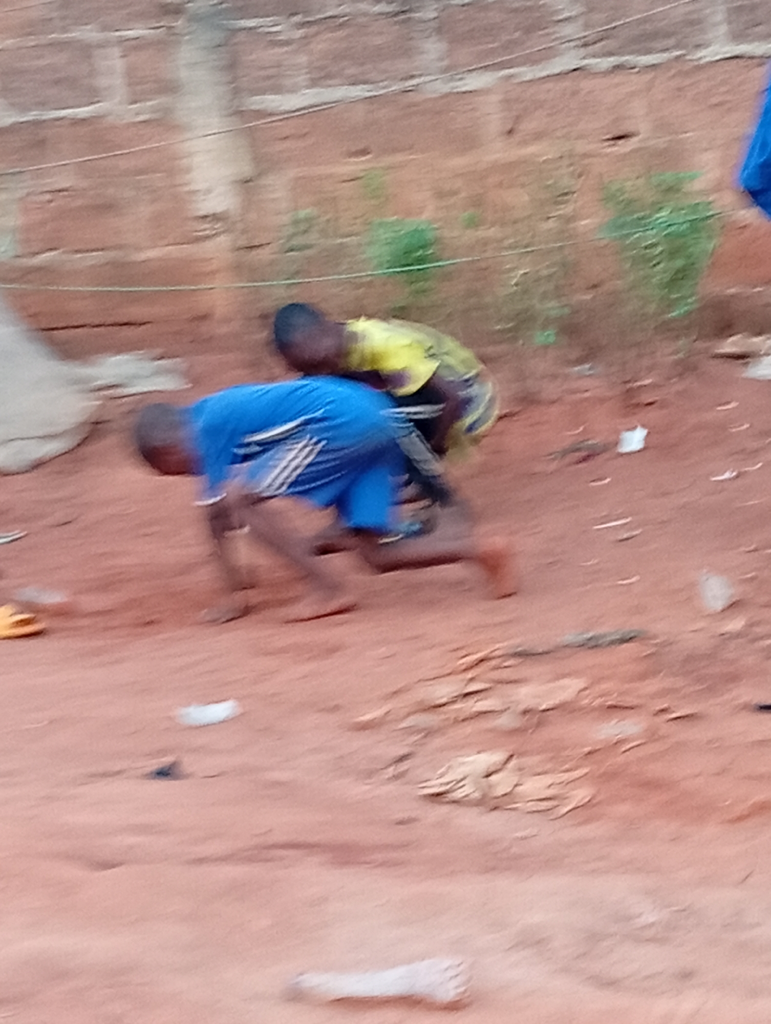 Two children play outdoors on reddish-brown soil. One child, wearing a blue shirt and shorts, is on all fours. The other child, wearing a yellow shirt, appears to be riding on the other’s back. The scene is in Ogidi, Nigeria, as indicated by the environment and subject actions. A brick wall is in the background, and sparse vegetation grows behind the children. Debris is scattered on the ground.