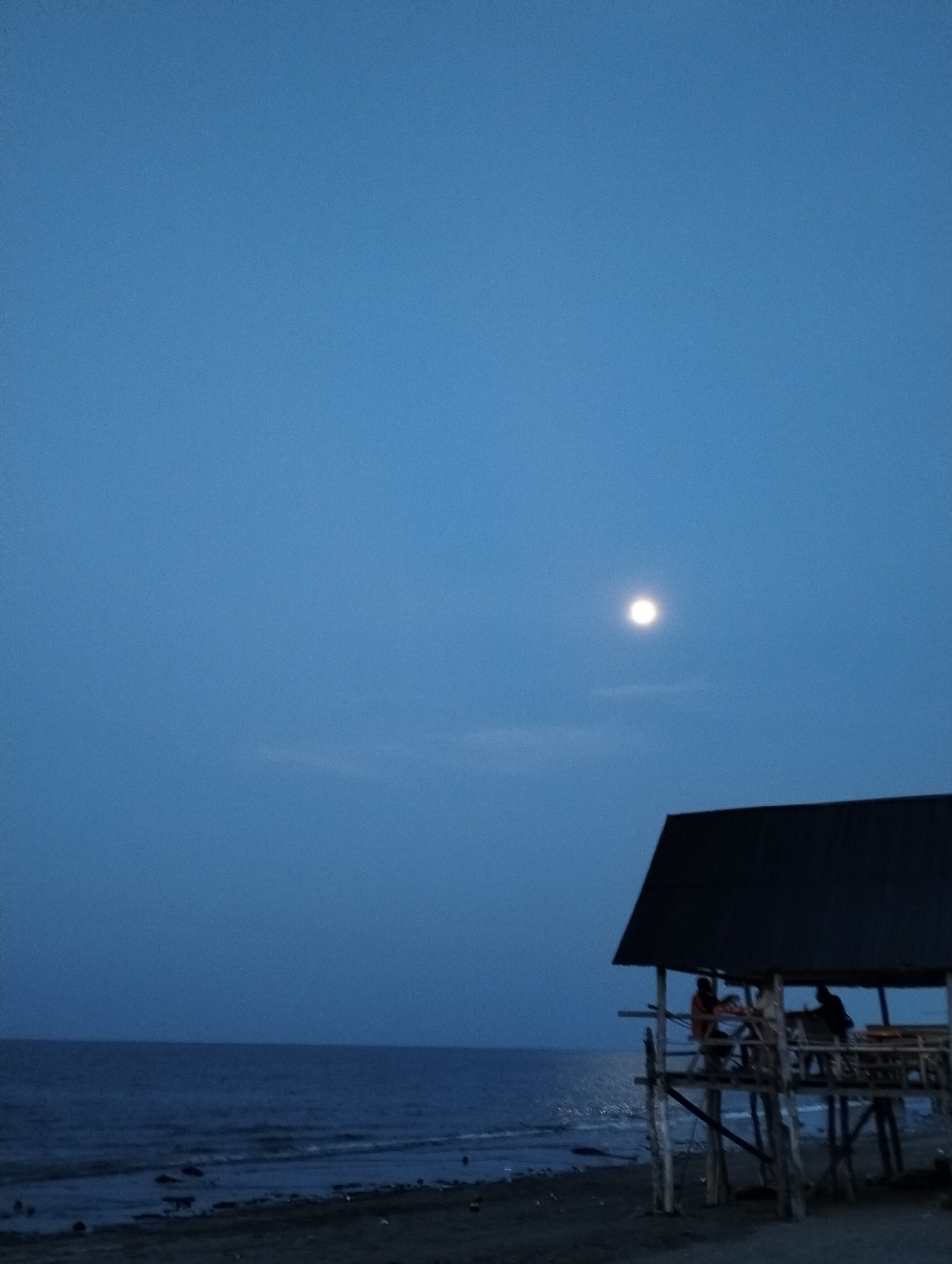 The image captures a serene coastal scene at dusk or early night in Lhokseumawe, Indonesia. The sky dominates the upper two-thirds of the frame, painted in a deep, consistent blue hue, suggesting a clear evening. A prominent, bright white moon shines in the upper right quadrant, casting a soft glow. Faint, wispy cloud formations are subtly visible below the moon and closer to the horizon line.
Below the sky, the vast expanse of the ocean appears dark blue, with gentle waves breaking onto a dark, rocky or sandy shoreline in the foreground. The water reflects some of the ambient light, particularly near the horizon.
On the right side of the image, a rustic, stilted wooden hut or pavilion stands near the water's edge. It features a dark, sloped roof and is constructed from wooden poles. Inside the open-sided structure, several people are visible, mostly silhouetted against the slightly brighter sea. At least three individuals can be discerned, with one person in the foreground wearing a light orange top, making them slightly more distinct. They appear to be seated, likely socializing or relaxing, enjoying the evening atmosphere by the sea.
The overall impression is one of calm and tranquility, with the bright moon serving as a natural focal point against the darkening sky and ocean. There is no visible text in the image.