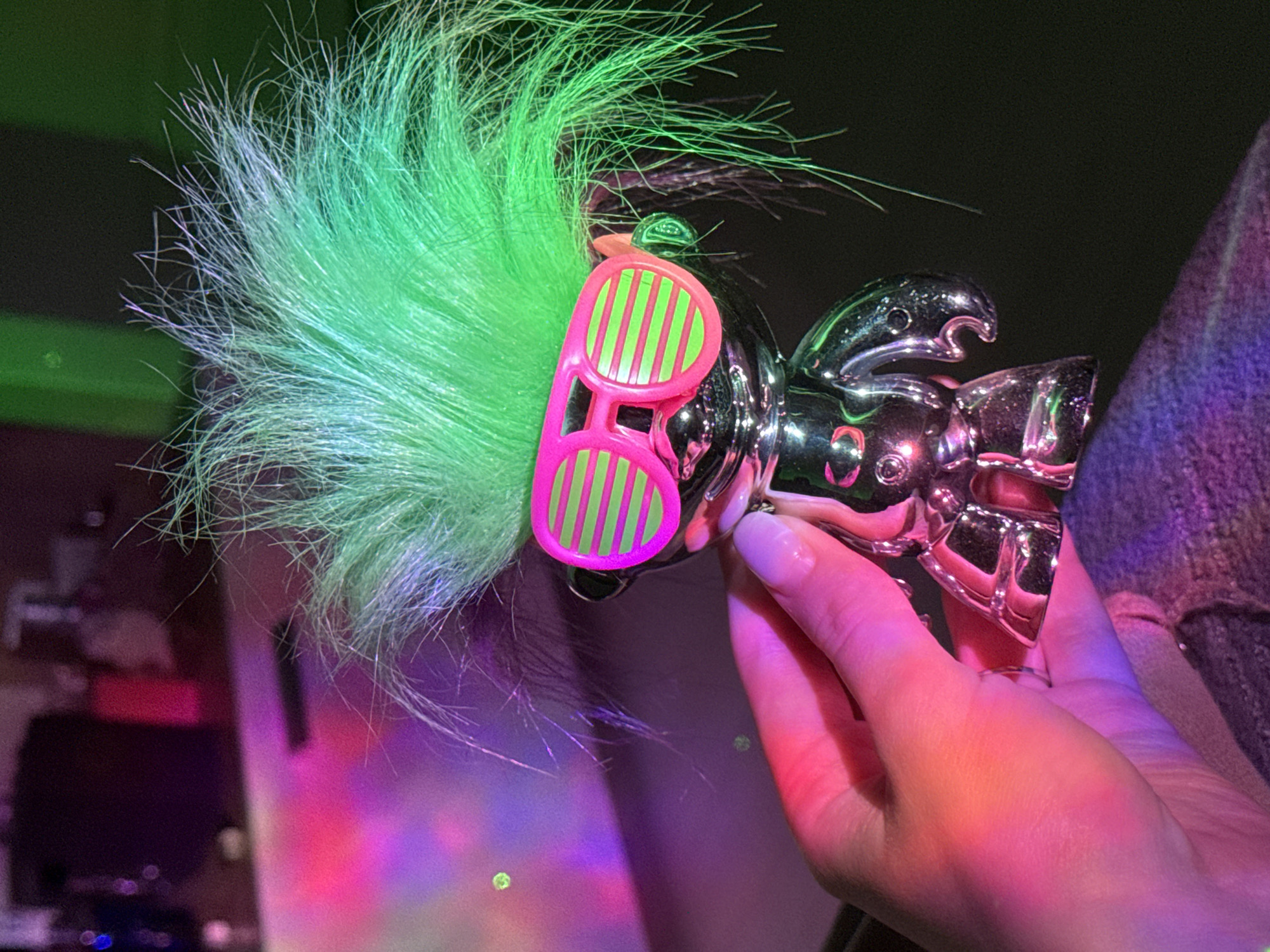 A hand holds a silver metal figurine with green hair and pink and green striped sunglasses. The background is dark and blurry. The location and city are unknown.