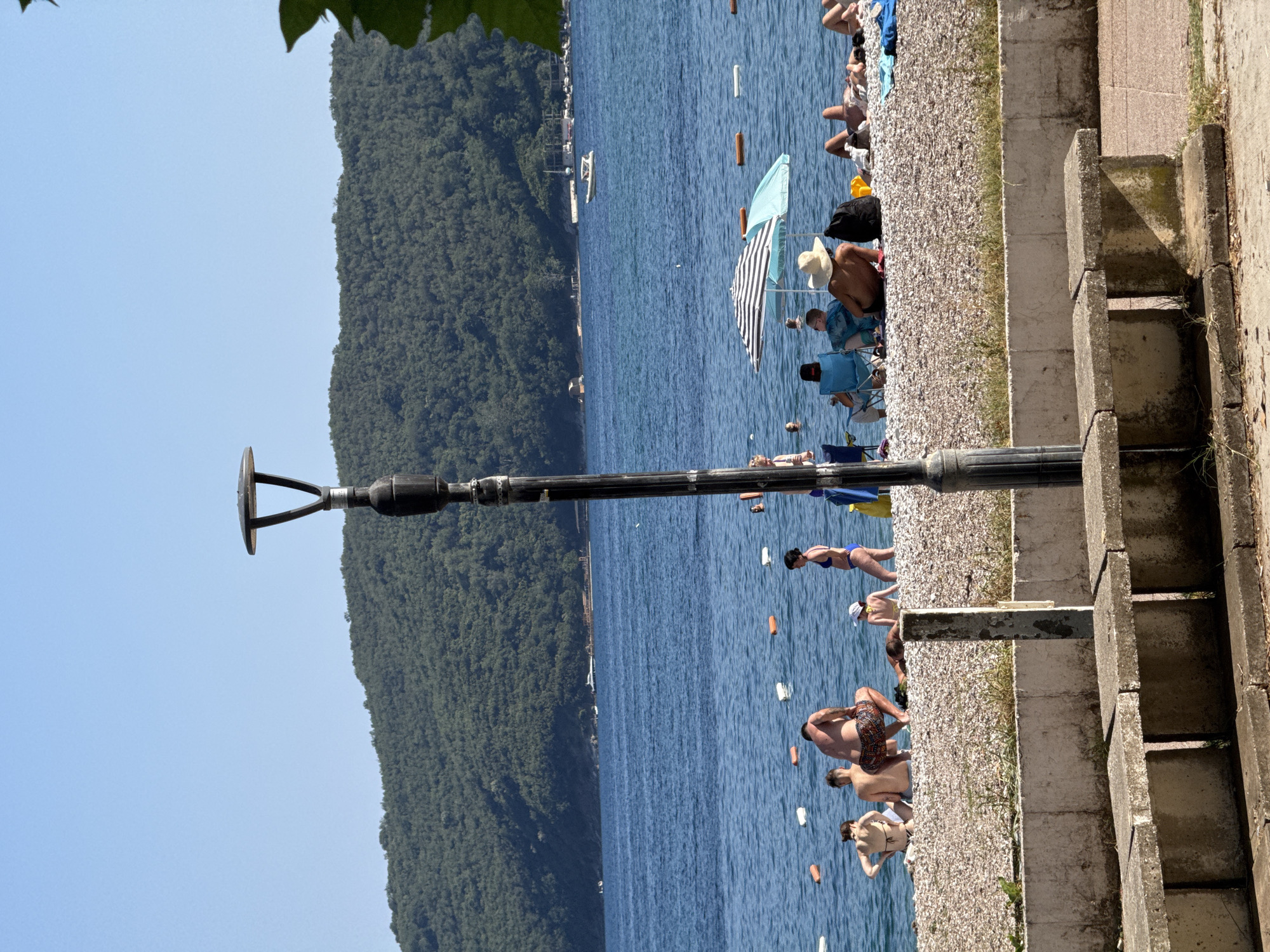 In Budva, Montenegro, a sunny day unfolds at a pebble beach. The foreground shows weathered concrete benches and a low wall. A tall lamppost stands prominently. Beyond, a bustling beach scene unfolds with numerous individuals enjoying the water and shore. Some are seated on the pebble beach, some are standing, and some appear to be swimming or wading in the calm, blue sea, marked by buoys and floating objects. Several beach umbrellas provide shade. In the background, a lush, green, forested hill rises, meeting a clear blue sky. The water is dotted with a few boats, hinting at activity beyond the immediate beach area.