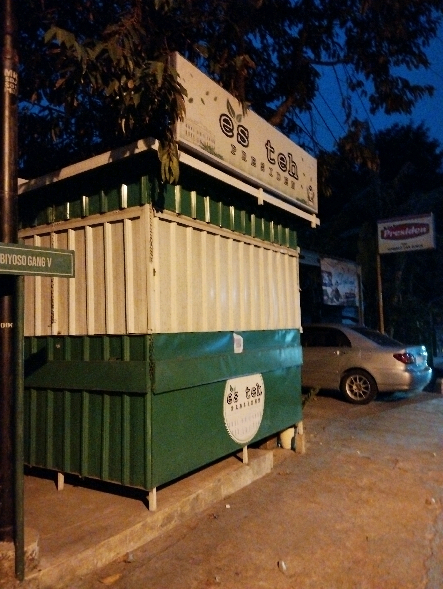 The image depicts a street scene at night in Salatiga, Indonesia. In the foreground, a green and white corrugated metal kiosk stands on a concrete surface, with a street sign that reads "BIYOSO GANG V" attached to a dark pole on the left. Above the kiosk, a sign displays "es teh PRESIDENT" in stylized lettering. Further back, a silver sedan is parked. A red and white "Presiden" sign is mounted on a pole to the right of the kiosk. The scene is illuminated by artificial lighting, creating shadows and highlighting the textures of the objects. Trees with dark foliage are visible in the background, against a twilight sky.