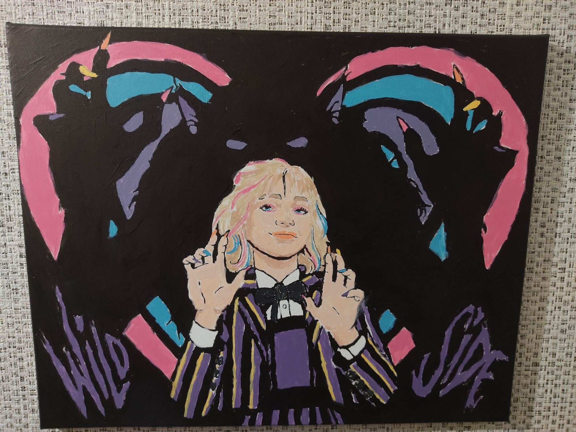 The image is a painting on canvas of a stylized portrait of a young woman with blonde hair streaked with pink and blue. She is wearing a black bow tie, a white collared shirt, and a purple and yellow striped jacket. Her hands are raised in front of her chest, with her fingers splayed in a gesture. The background is black and features abstract shapes and figures in pink, teal, and purple. There are two large, curved pink shapes behind the woman's head, framing her. On the bottom left and right of the painting, there are stylized purple letters that appear to read "WILD" and "SIDE". The painting is displayed on a textured surface, possibly a wall or table, with a faint cross-hatch pattern visible. The painting is located in the city of Rozsoshentsi, Ukraine.