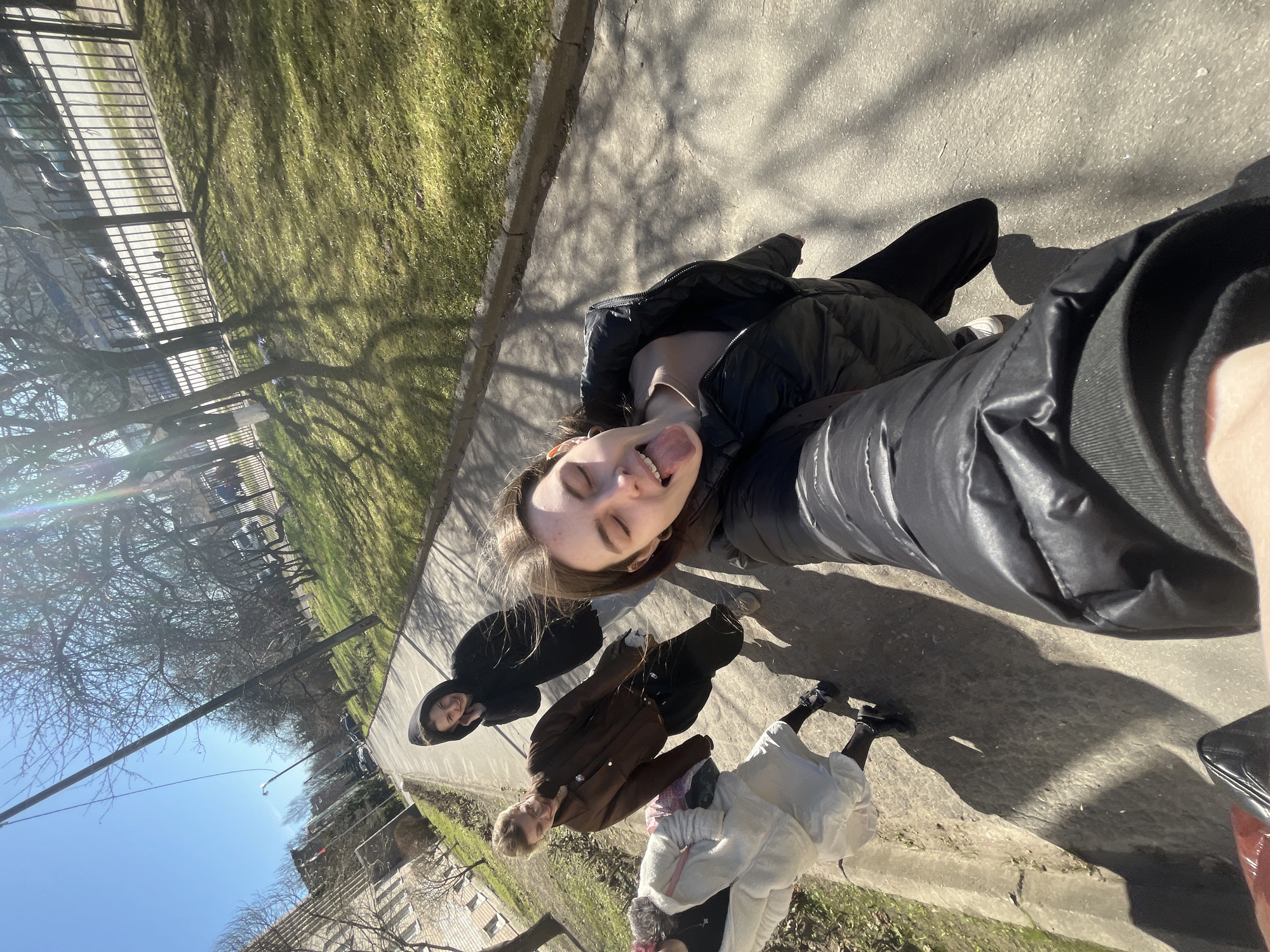A selfie taken outdoors in Saint Petersburg, Russia, featuring four individuals on a paved surface alongside a grassy area with trees. The primary subject, a young woman wearing a black puffer jacket, is holding the camera and sticking out her tongue. Behind her are three other individuals, one in a black hooded jacket, one in a brown jacket, and another in a white dress. The background includes a metal fence, buildings, bare trees, and a blue sky. Sunlight creates shadows on the ground.
