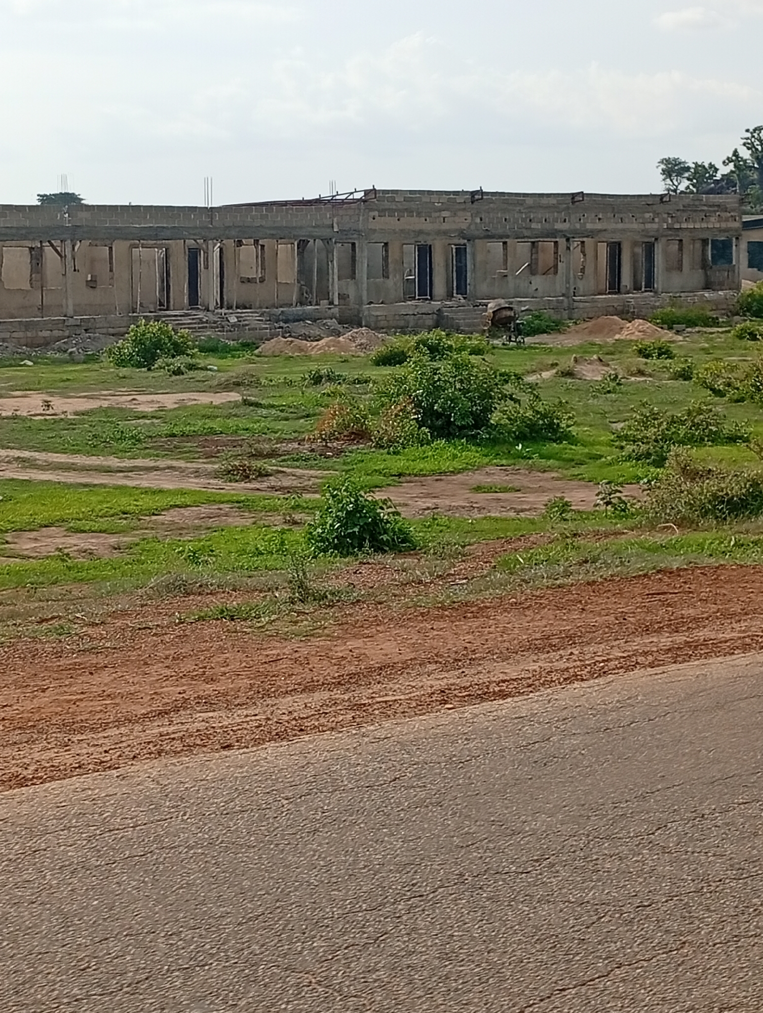 The image shows an exterior view of a long, rectangular building under construction in Shuwa, Nigeria. The building appears to be a single-story structure with an open roof and partially completed walls made of concrete blocks. Numerous doorways and window openings are visible along the facade, some with exposed wooden framing. The surrounding environment consists of a grassy, unkempt field with patches of dirt and scattered vegetation. In the foreground, a dirt road or path runs diagonally across the lower portion of the image, leading to a paved road. The sky is overcast with a few visible clouds. There are no people or vehicles visible in the scene.