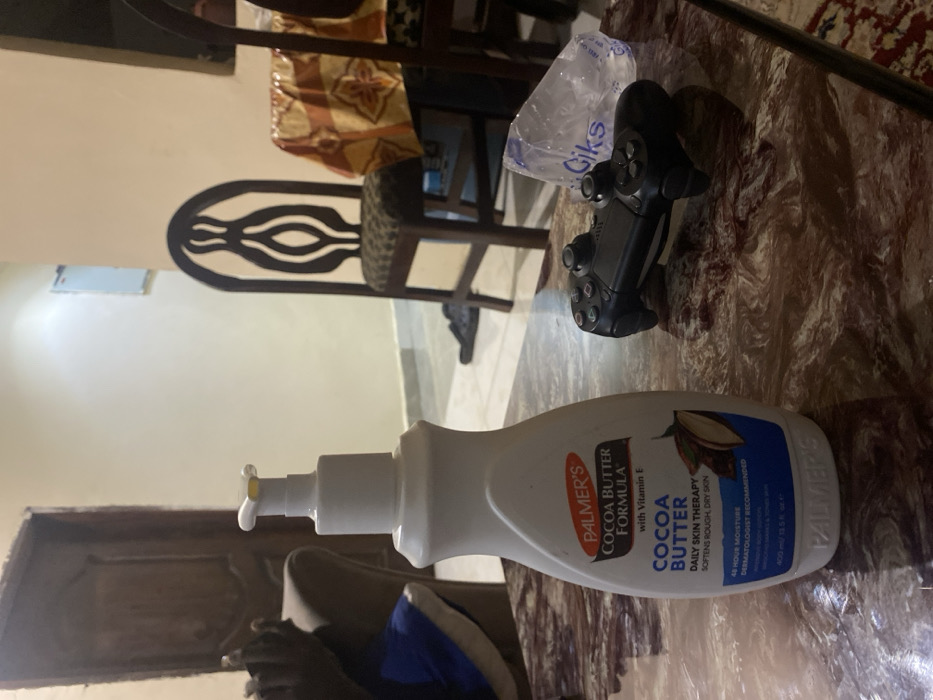 The image displays an indoor scene, likely a living or dining area, in Lakka, Sierra Leone. The primary focus is a white pump bottle of Palmer's Cocoa Butter Formula lotion, standing upright on a table with a marbled surface in the foreground. To the right of the lotion bottle, a black PlayStation controller is visible, alongside a clear, crumpled plastic bag with the word "Giks" printed on it.
In the background, various pieces of dark wooden furniture can be seen, including chairs. One chair's backrest features a distinctive, ornate design with elongated, eye-shaped cutouts. The walls are plain and light-colored, suggesting a simple interior decor. The floor also appears to be a light shade. A piece of upholstered furniture, possibly a sofa or armchair, is partially visible in the lower-left corner. No specific activity or event is taking place; the scene captures a static arrangement of everyday objects.
Notable text on the Palmer's lotion bottle includes "COCOA BUTTER FORMULA with Vitamin E", "COCOA BUTTER DAILY SKIN THERAPY SOFTENS ROUGH, DRY SKIN", "48 HOUR MOISTURE", "DERMATOLOGIST RECOMMENDED", and "400 ml / 13.5 fl. oz.". The word "Giks" is also clearly legible on the plastic bag. The lighting is consistent with an indoor setting, but does not indicate a specific time of day.