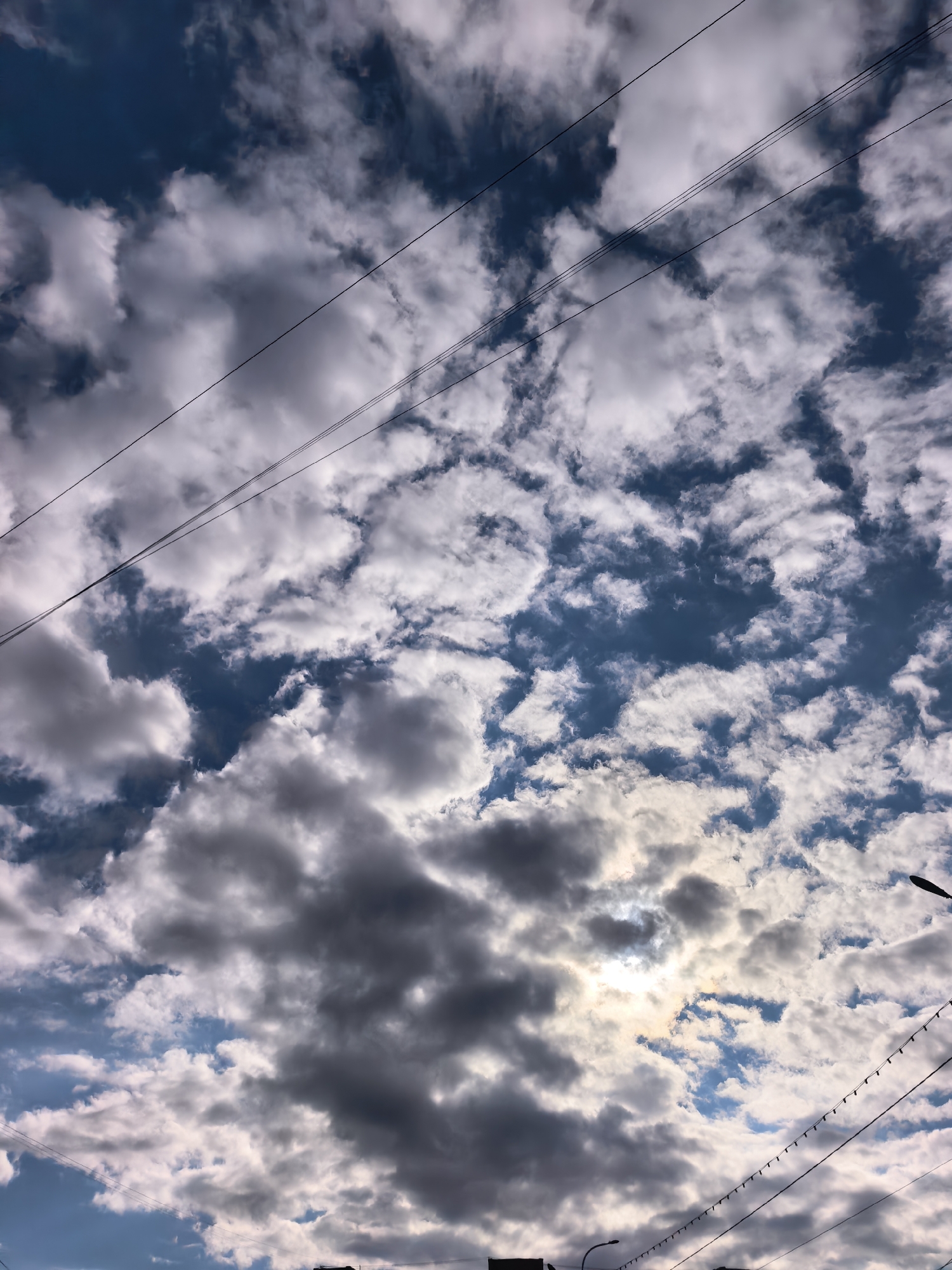 The image captures a sky filled with cumulus clouds, some appearing dark grey and others lighter, against a backdrop of blue. Sunlight penetrates through the clouds, creating a bright area. Several overhead power lines intersect the view, stretching across the sky. In the bottom of the image, silhouetted rooftops and streetlights suggest an urban setting. While the location is specified as Sterlitamak, Russia, there are no distinctive elements confirming this location; the scene is common to many urban areas. The scene lacks people and action, and the focus is on the atmospheric conditions and urban infrastructure.