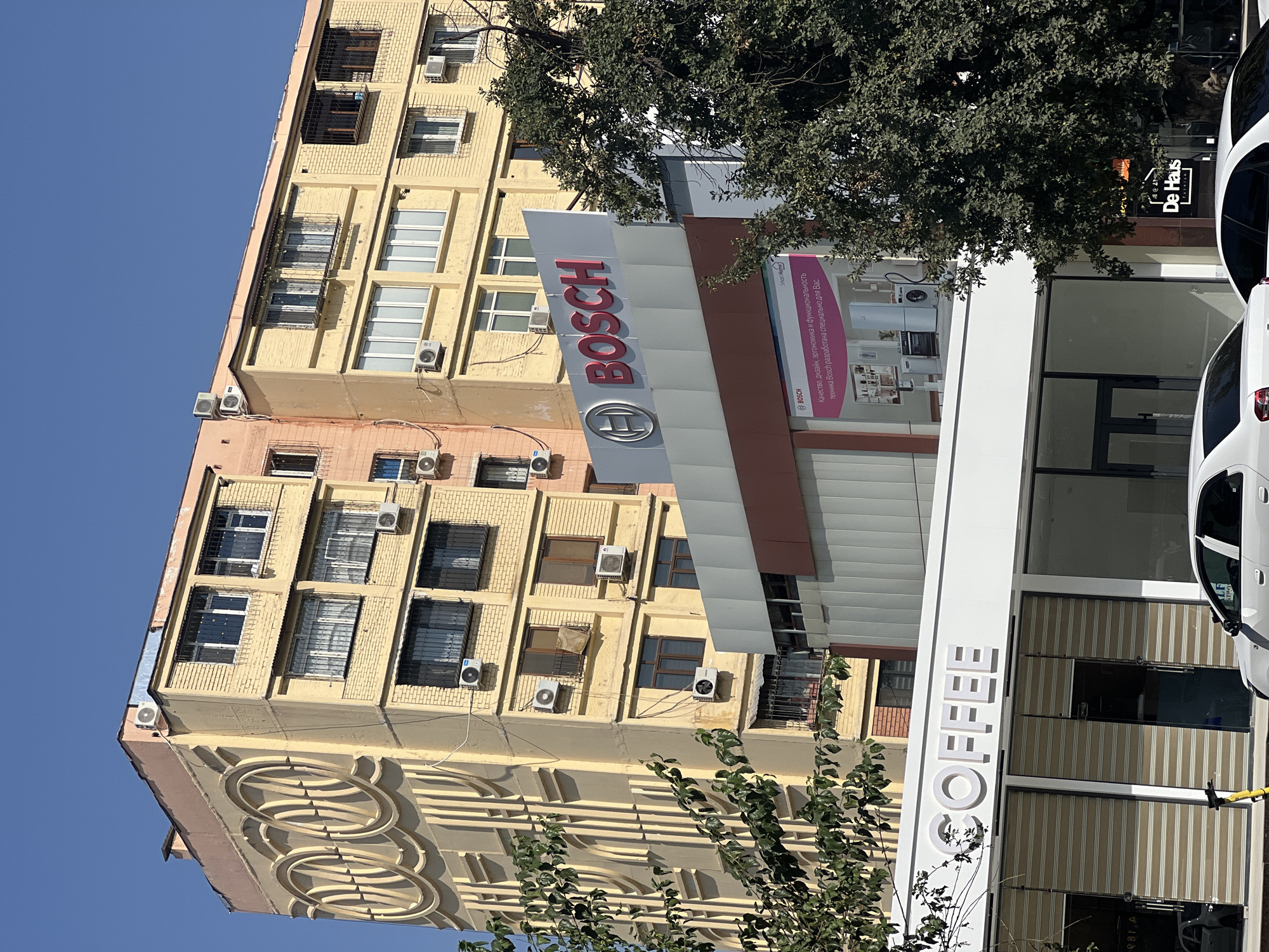 The image captures a street view in Tashkent, Uzbekistan, on a clear, sunny day. In the foreground, a modern building features a white facade with the word "COFFEE" prominently displayed in large, white, three-dimensional letters. Below this signage, a storefront with closed roll-up shutters and large glass windows is visible, with a yellow electric scooter parked to the left. To the right of the coffee shop, another building displays a sign for "De Haus Premium."
Above the coffee shop, a multi-story building with a light yellow brick facade is partially visible. This building has numerous windows, many fitted with security bars, and several air conditioning units mounted on its exterior. A prominent red Bosch logo is displayed on a separate structure in front of this building, accompanied by a large advertisement showcasing Bosch appliances. The background is dominated by a tall, pale yellow apartment building with unique circular decorative elements on its left side, extending upwards and featuring multiple rows of windows. The sky is a clear, bright blue.