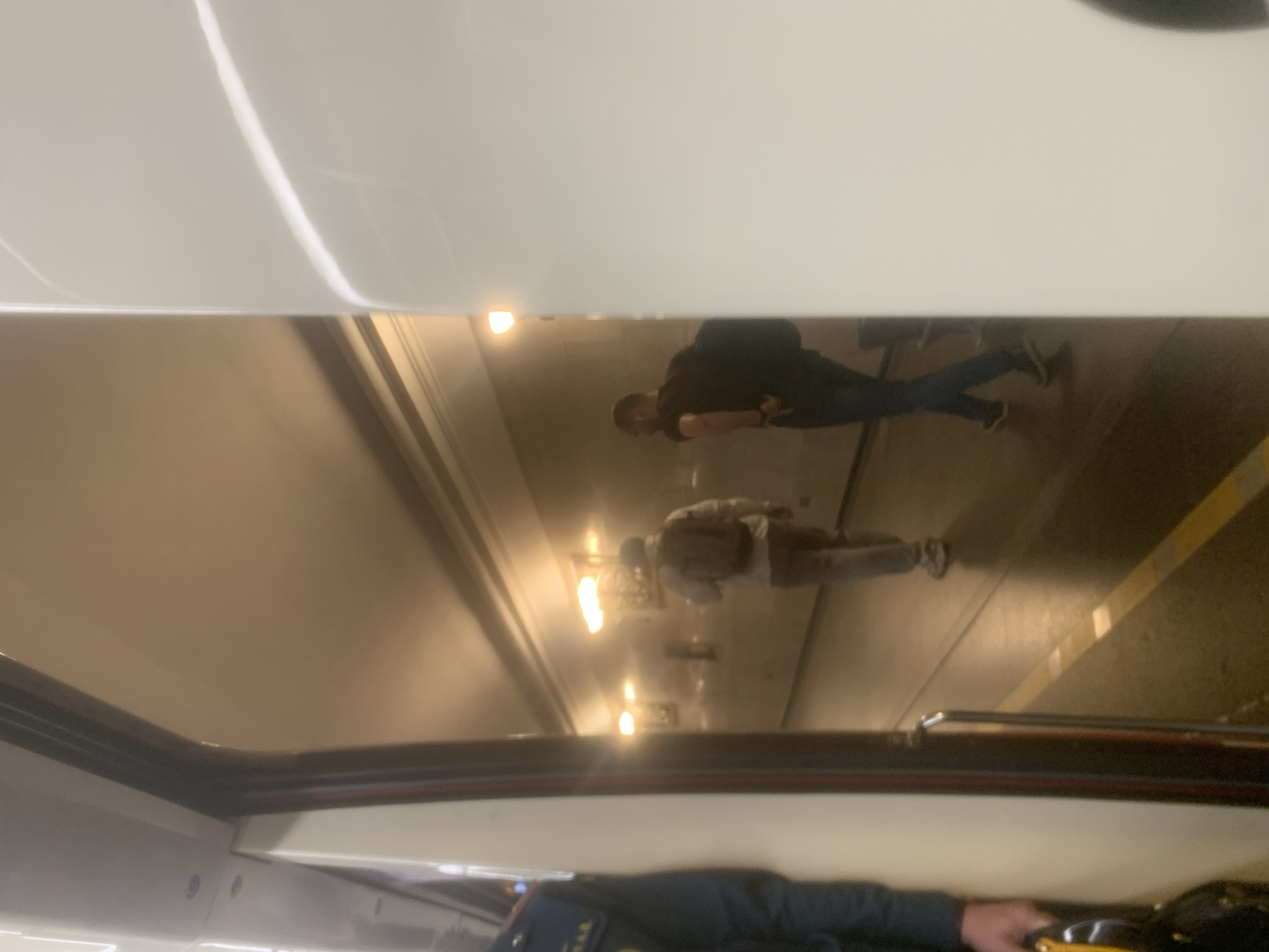 The image, appearing to be rotated 90 degrees clockwise, captures a scene from inside a vehicle, likely a train or metro car, looking out a window. It simultaneously displays a reflection of the vehicle's interior and a view of an external environment.
The upper half of the image depicts what appears to be an underground tunnel or metro station platform. Two men are visible on the platform. The man on the left wears a dark top, dark pants, and a backpack, and seems to be looking downwards. To the right and slightly further back, a second man is dressed in a lighter-colored top and pants, also carrying a bag or backpack, and appears to be standing or walking. The background reveals a long, curved tunnel illuminated by multiple bright, warm-toned light fixtures along its walls. A yellow line is visible on the platform floor towards the right, likely marking a safety boundary.
The lower half of the image shows the interior of the vehicle and its reflection. On the bottom left, a blurry reflection of the vehicle's white or light-colored ceiling is visible. On the bottom right, the lower part of a window frame is seen, along with the arm and hand of a person inside the vehicle. This individual is wearing a dark blue uniform-style jacket, and a patch with extremely indistinct, blurry Cyrillic letters is visible on their sleeve. Their hand holds a yellow object, possibly a handle or part of a personal item.
The artificial lighting and subterranean setting strongly suggest an underground metro system, consistent with the provided location of Saint Petersburg, Russia. The blurriness of some elements might indicate movement of the vehicle or low light conditions. No clearly legible text is present.