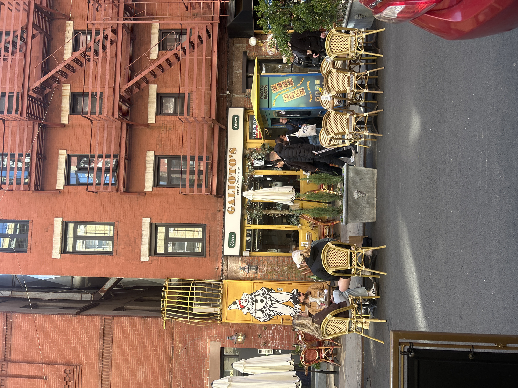 This image depicts a street scene in New York City, United States of America, featuring a deli named Galioto's. The deli, characterized by a yellow exterior and signage, has an outdoor seating area with chairs arranged along the sidewalk. The building has a red brick facade with a prominent fire escape. The street scene also includes pedestrians, some of whom are seated at the deli's outdoor tables. A photo booth is placed near the deli entrance. A red car is partially visible at the top right corner of the frame.