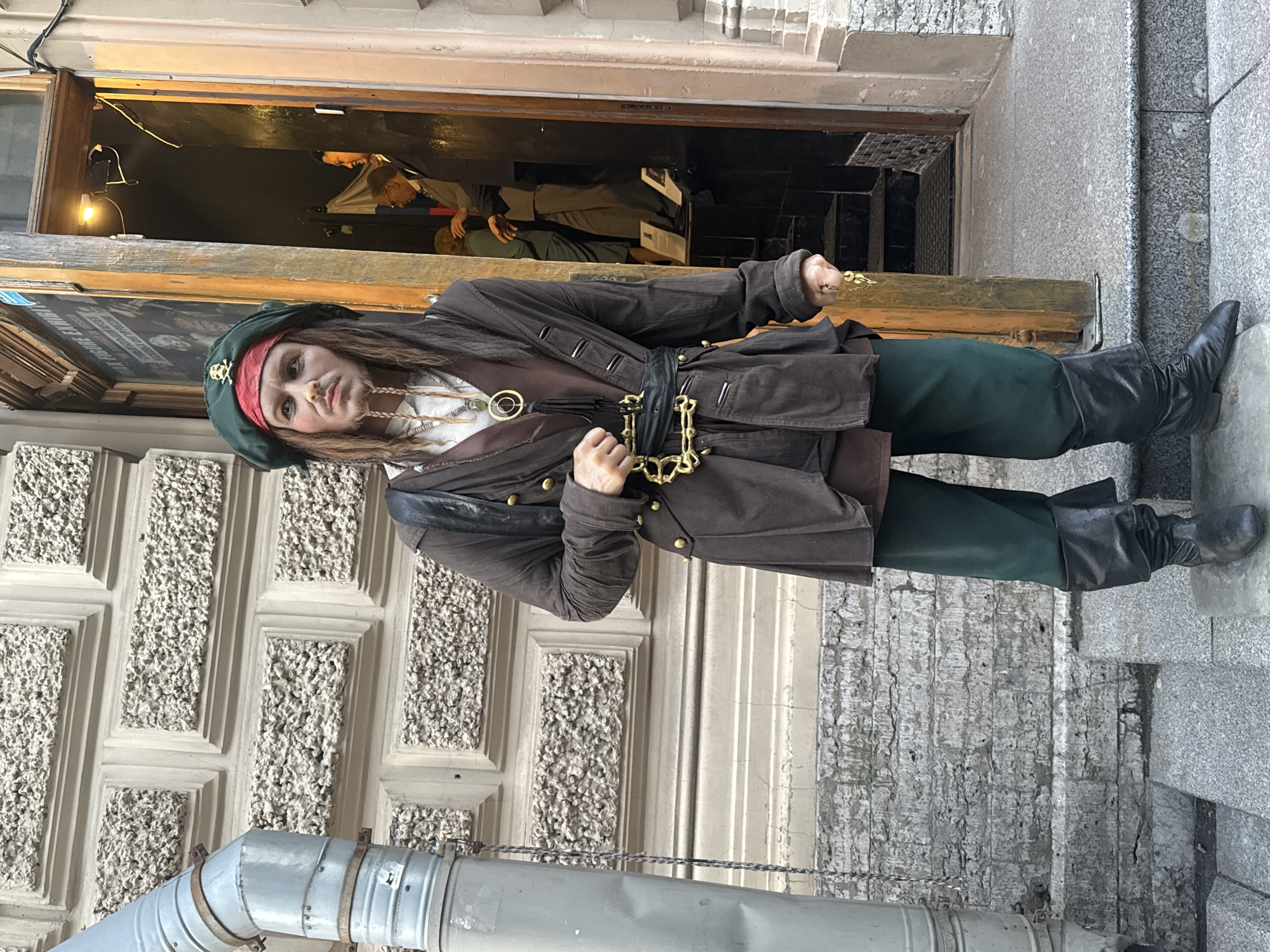 The image shows a person dressed as Captain Jack Sparrow, a character from the Pirates of the Caribbean film series. The subject is standing in front of an open doorway that appears to lead into a shop or establishment. The person is wearing a green tricorne hat with a skull and crossbones emblem, a red bandana, a brown coat with gold buttons, a white shirt, a dark sash around the waist, green pants, and black leather boots. A gold chain necklace with a circular pendant is visible around the neck.
The background shows the interior of the doorway, with a dimly lit space and what appears to be mannequins or other figures. The exterior features a stone facade with decorative paneling and a large metal pipe running along the bottom of the frame. The overall impression is that this is a street scene in a city, likely a tourist area given the costume. The lighting suggests it might be daytime, though the interior of the doorway is dark. The person's expression is somewhat serious and characteristic of the character they are portraying. The setting appears to be Saint Petersburg, Russia, as indicated by the location context. There is no visible text in the image.