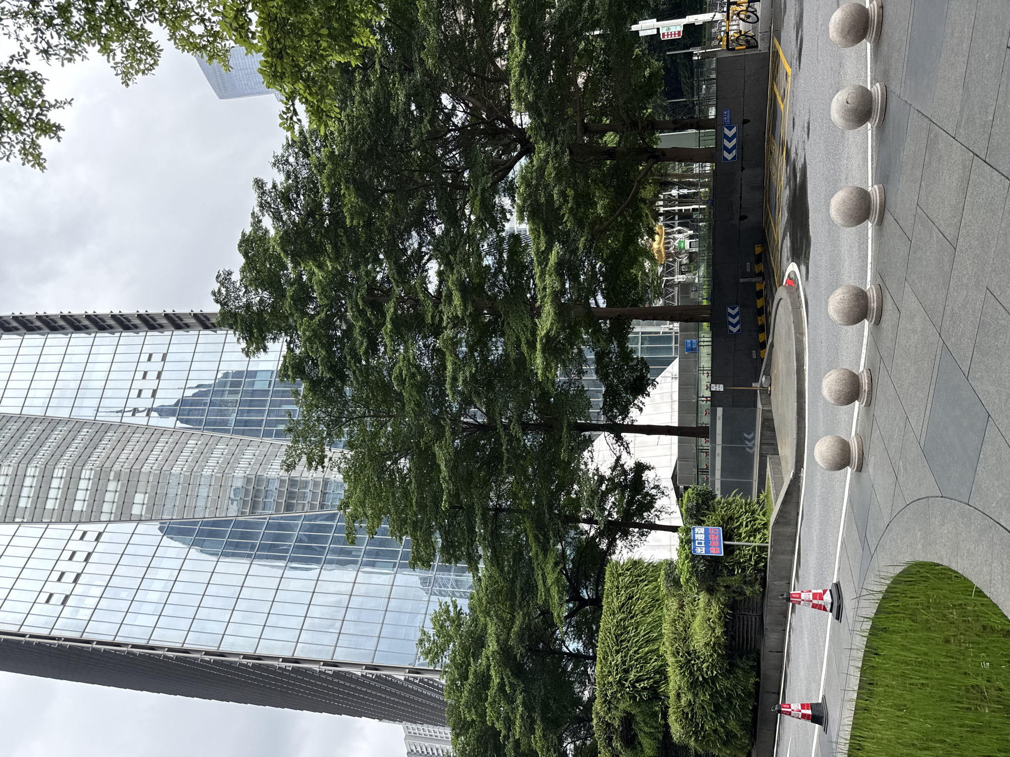 The image depicts an outdoor scene in Tianhe District, China, dominated by a modern glass skyscraper. The skyscraper has a faceted design, with its mirrored surface reflecting the sky and adjacent buildings. In the foreground, a well-maintained landscape features lush green bushes and trees, some of which partially obscure the lower levels of the skyscraper. A paved walkway with a curved edge is visible, bordered by a decorative arrangement of round, light-colored stone bollards. Traffic cones, one red and white and another orange and white, are placed on the road. A blue sign with Chinese characters is mounted on a pole in front of a dense green hedge. Further back, a dark gray retaining wall is visible, marked with blue directional arrows. A yellow bicycle is parked to the right, near a parking garage entrance. The sky is overcast.