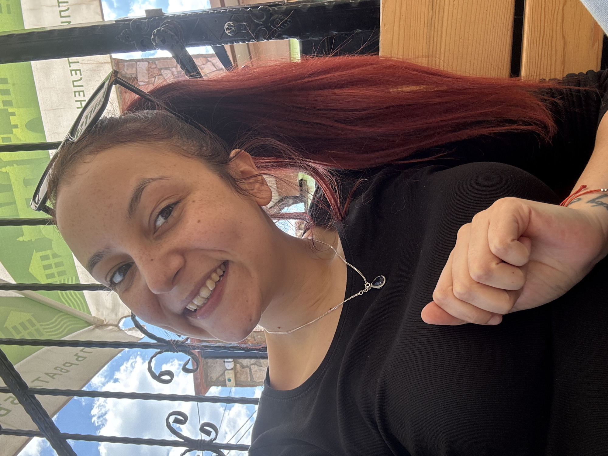 A smiling woman is visible in this selfie-style image. She has red hair and is wearing a black shirt and a silver necklace with a dark pendant. Glasses are perched on her head. Her right hand is clenched into a fist. The background reveals a wrought iron structure, a green and white awning with Cyrillic script, and a partly cloudy sky. The woman appears to be sitting at a cafe. This location is in Dobrich, Bulgaria.