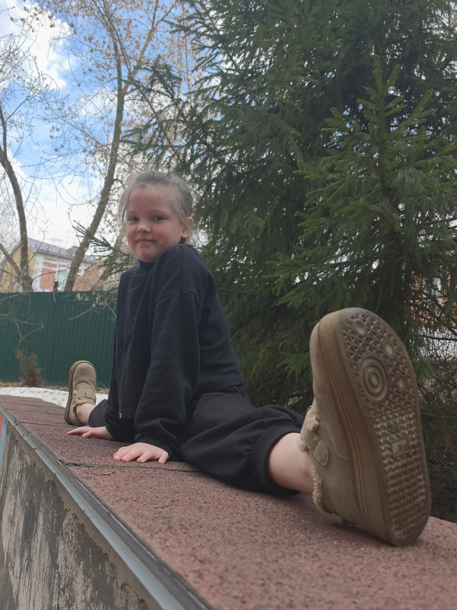A young girl is performing a split on a low concrete wall. She is wearing a black sweatshirt and pants, and beige sneakers. The girl is looking at the camera with a slight smile. The background includes a green fence, a house, trees, and a blue sky with clouds. The photograph was taken in Nizhny Novgorod, Russia.