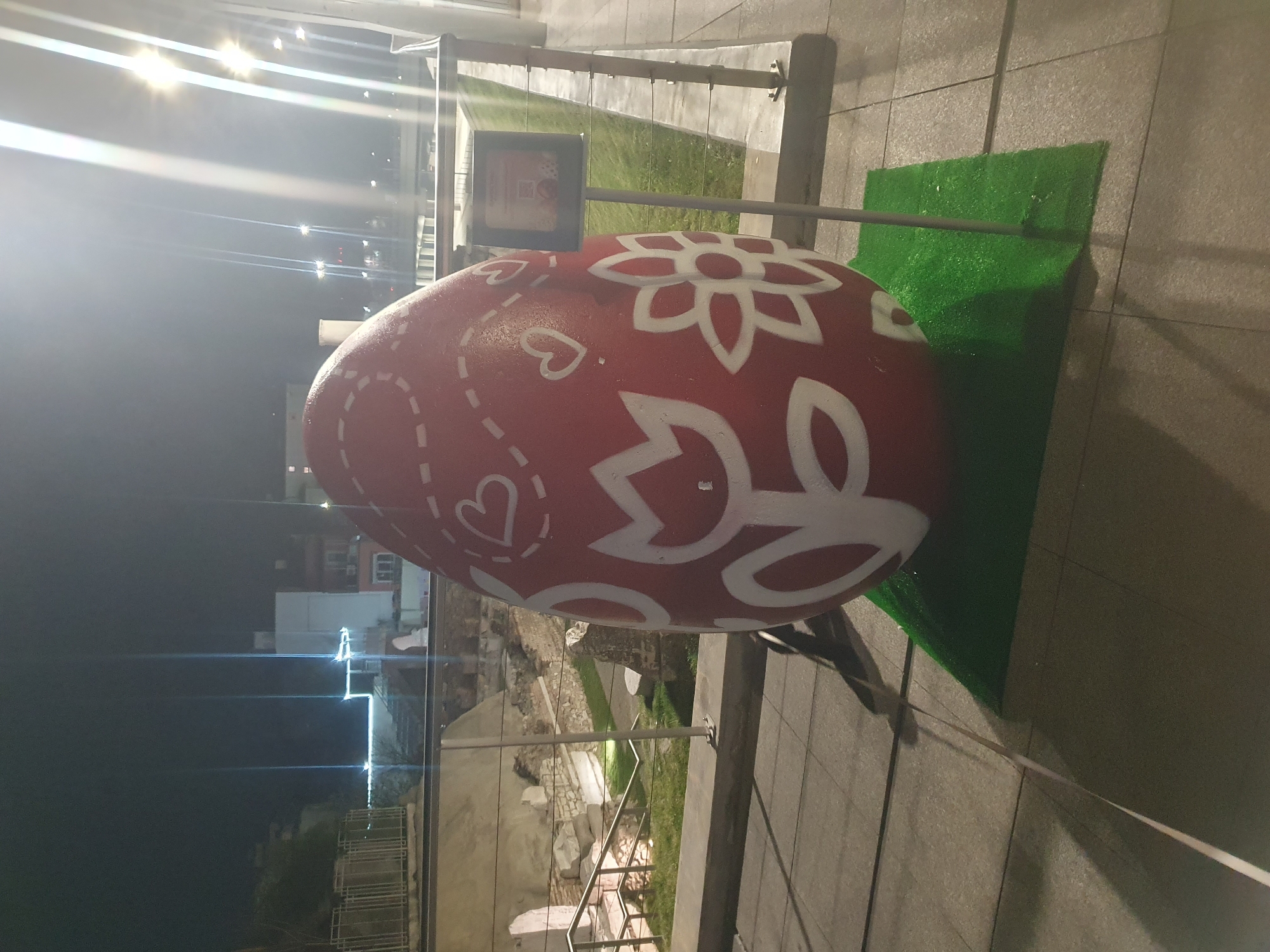 A large decorative red Easter egg with white flower and heart designs sits on a green patch on a gray paved surface. A metal support structure is attached to the egg. Behind the egg and support, ancient ruins can be seen, with a modern building in the background. The time of day appears to be night, with bright artificial lighting. The location is likely the center of Plovdiv, Bulgaria, near the Roman Stadium ruins.