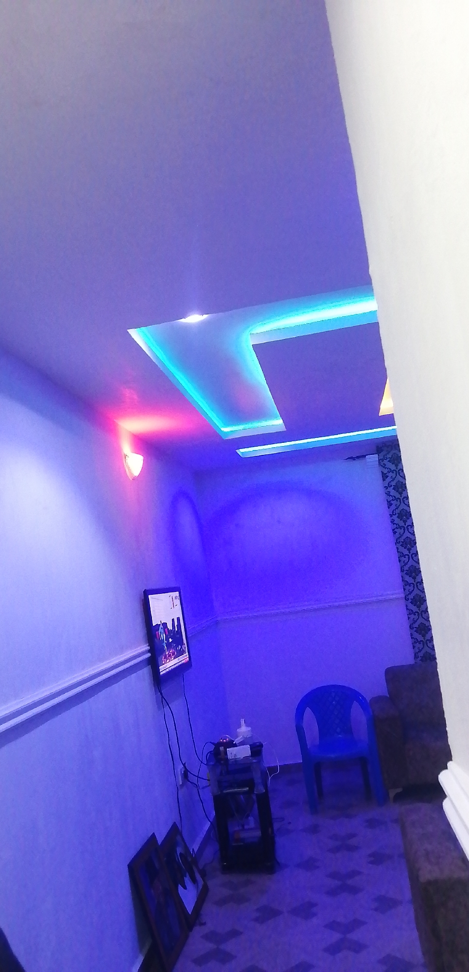 The image depicts an interior scene, likely a living room, bathed in the purple and blue glow of ambient lighting. The ceiling features modern recessed lighting with blue LED strips accentuating geometric contours. A wall-mounted flat-screen television displays an image, while a small, dark stand sits below it, holding various electronic devices and a bottle of liquid. To the right of the stand, a bright blue plastic chair is positioned. In the background, a patterned curtain and a dark-colored sofa are visible. On the floor, two framed pictures lean against the wall near the television. The overall impression is one of a contemporary and vibrantly lit space.