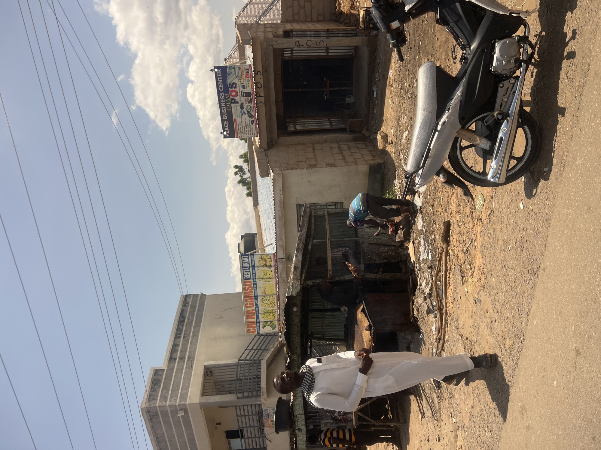 The image captures a sunny street scene in Chum, Nigeria, identified by a sign displaying "PEACE BUSINESS CENTER CHUM." The sky is clear blue with scattered white cumulus clouds, and strong sunlight casts distinct shadows, indicating midday conditions.
In the immediate foreground, a paved road surface transitions into an unpaved, dusty, gravelly shoulder littered with sticks, plastic, and various debris. A dark-colored motorcycle with a grey seat and chrome exhaust pipe is parked on the right side of this transition, with some parts wrapped in plastic.
On the left side of the midground, a man in a long white robe (thawb/jellabiya) and a black-and-white checkered scarf stands facing slightly right. Behind him and to his left, a man in a striped yellow and dark shirt is partially visible near a wooden table. Directly behind them is a makeshift open-air vendor stall with a corrugated metal roof, where two individuals are active. One man in dark clothing is visible handling what appears to be cuts of meat on a surface, while another man in a blue and white striped shirt is bending over near the ground to the right of the stall. A small yellow generator is visible near this bending figure.
The background features several buildings. On the left, a two-story building with a light-colored facade features dark horizontal stripes on its upper section. It has a balcony with metal railings and a barred window. A prominent sign for "CHI KA GAMSU RESTAURANT" advertises food items such as "JOLLOF & WHITE RICE," "TUWO," "CHIPS," "NOODLES & EGG," and "PEPPEED SOUP." Below this, a partial sign reads "JAUKA'U CLOTHING." A black water tank rests on the roof of this building. On the right, a single-story concrete block building houses the "PEACE BUSINESS CENTER CHUM" and "P.O.S" (Point of Sale) services. Its sign lists multiple services including "Mtn, Glo, Airtel, 9mobile, Data Subscriptions, Digital Passwording, School Fees Payment, Phone Accessories, Recharge Cards," and "ETC." The building's wide entrance is open, revealing a dark interior. "P.O.S" is also painted vertically on the exterior wall beside the entrance.
Multiple thin, dark power lines traverse the upper portion of the sky, extending across the scene. Sparse green vegetation is visible in the distant background on the right.