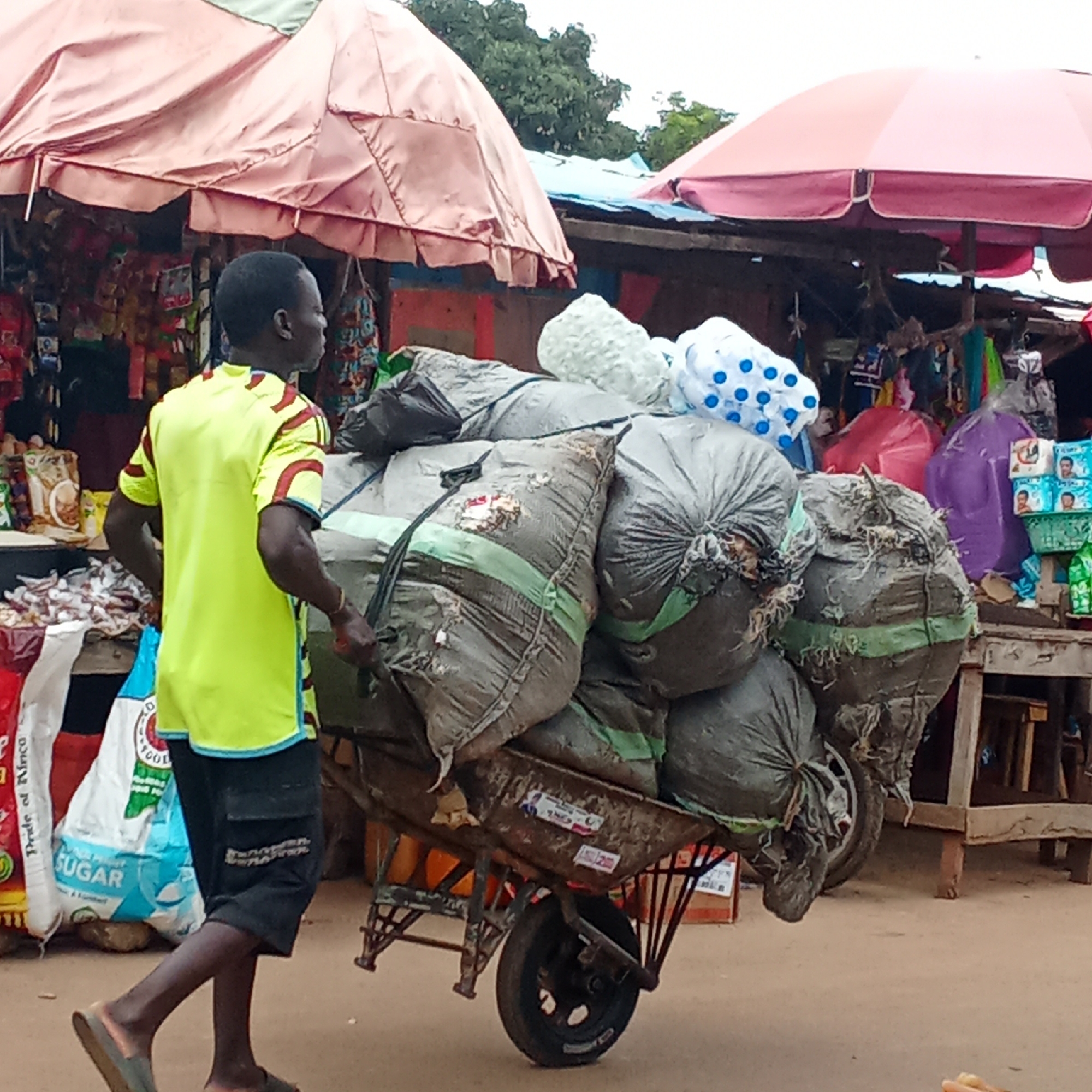 In Akwanga, Nigeria, a man in a yellow and maroon t-shirt and dark shorts pushes a heavily laden wheelbarrow. The wheelbarrow is filled with gray sacks tied with green straps and white bags. The scene is a market, indicated by stalls covered with red and pink umbrellas and stocked with various goods. A bag of sugar with "Pride of Africa" printed on it is visible near the wheelbarrow.