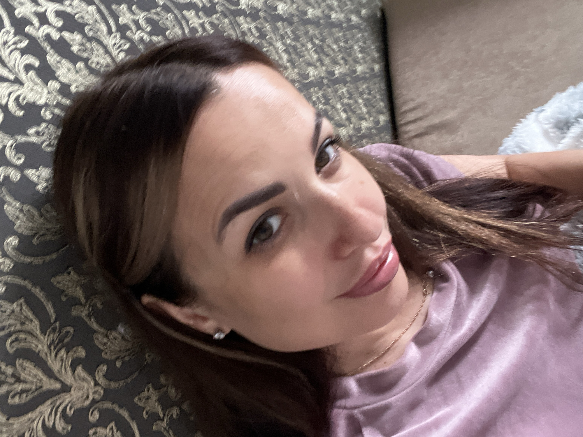 The image shows a close-up, high-angle selfie of a woman with brown hair, wearing a pink t-shirt and a necklace. She is lying down on a textured surface, possibly a couch, with a patterned wallpaper in the background. Her head is tilted, and she is looking towards the camera with a slight smile. A silver earring is visible on her left ear. The lighting is soft, creating a warm glow on her skin.