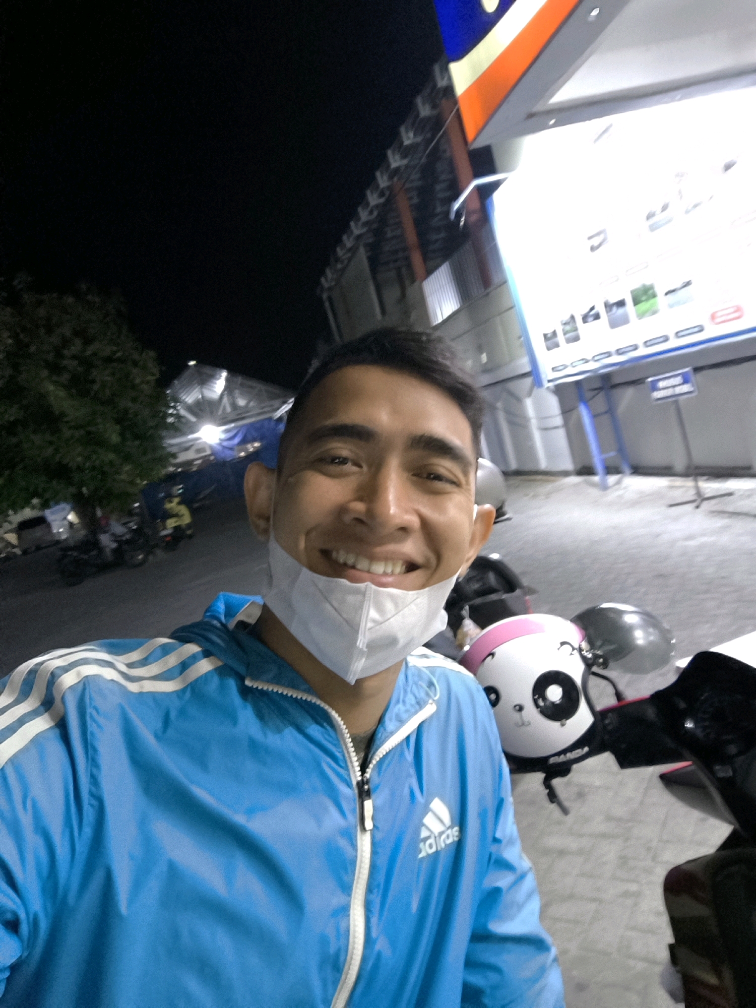 In a nighttime scene in Limboto, Indonesia, a young man wearing a blue Adidas jacket with white stripes and a white face mask smiles for a selfie. He is positioned in the foreground, with his face partially obscured by the mask, revealing a bright smile and dark hair. Behind him, a white and pink helmet with a panda design is visible on a motorcycle. The background features a brightly lit building with a sign and an illuminated display, likely a convenience store or gas station, common in urban Indonesian settings. A tree with dense foliage is also present, along with streetlights and other motorcycles in the distance, suggesting a lively, commercial area.