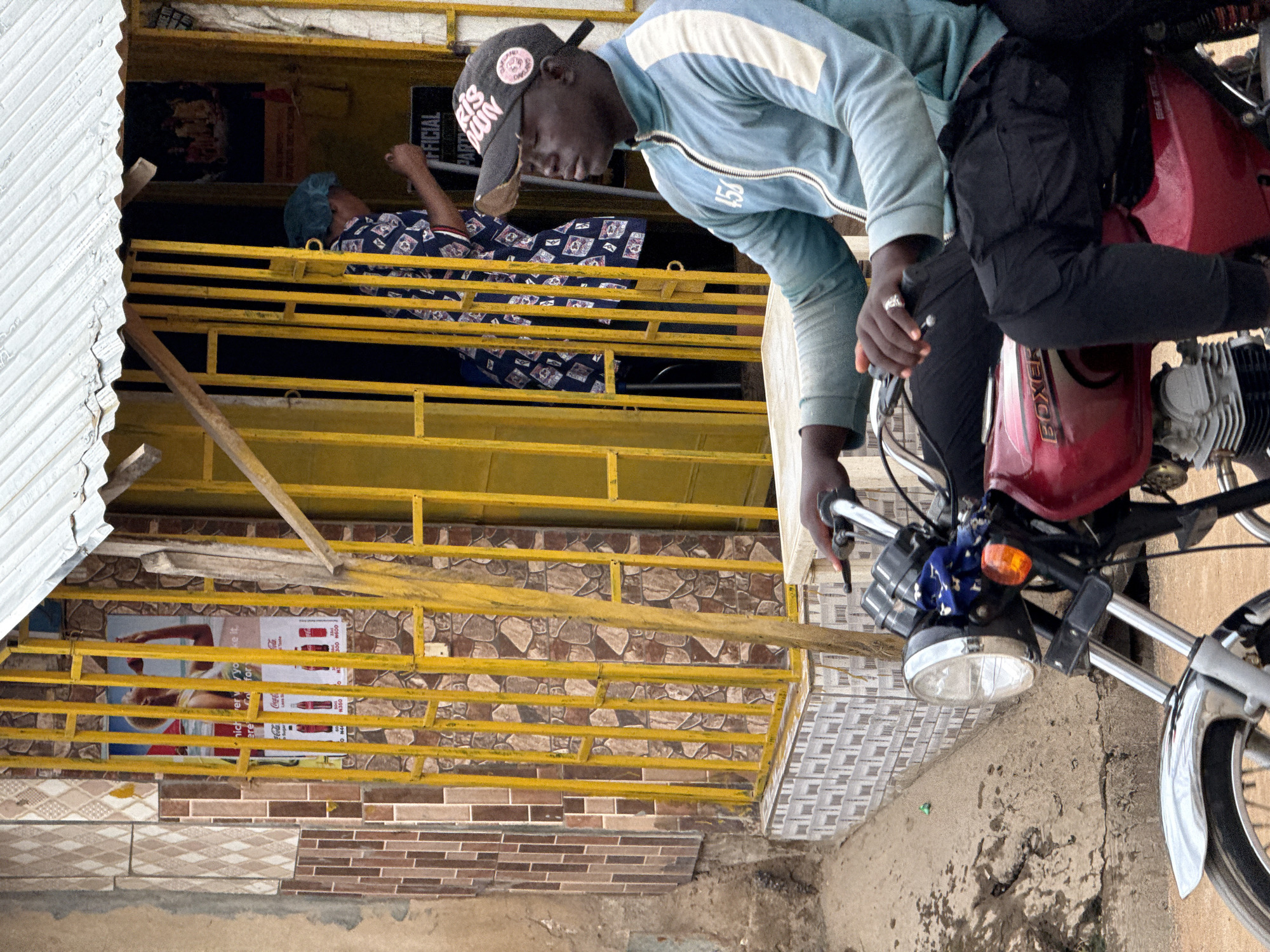 This image, set in Abuja, Nigeria, captures a vibrant street or market scene with two individuals and a motorcycle. The perspective is from a slightly elevated position, looking down.
In the foreground, a dark-skinned man is prominently featured, leaning over a red motorcycle. He wears a light blue zipped jacket with white stripes on the sleeves and a dark baseball cap with some white text, possibly reading "RIS MAIN". A silver ring is visible on his left ring finger. His gaze is directed downwards, suggesting he might be inspecting the motorcycle or looking at something on the ground. The motorcycle's fuel tank clearly displays the brand name "BOXER" in white lettering. The front part of the motorcycle, including the headlight and chrome fender, is visible, indicating it's a common commuter bike.
Behind the man and slightly above, there is a yellow metal balcony structure. On this balcony, another person is partially visible, lying down or resting. They are wearing a patterned dark blue and white shirt and a blue head covering.
The background reveals more architectural details: yellow metal railings form part of the building's structure. Various tiled surfaces are visible, including intricate brown patterned tiles on the wall behind the balcony and a mix of brown brick-like tiles and geometric patterned tiles on the lower walls. There are also posters affixed to the walls; one dark poster has the word "OFFICIAL" visible, while another features Coca-Cola bottles with accompanying text like "Coca-Cola" and price indications such as "300 DOLLARS" (likely Naira, given the context) and "N500". The left side of the image shows corrugated metal siding, typically used for roofing or partitioning.
The scene is brightly lit, suggesting it is daytime. The weather appears clear. The overall impression is one of everyday activity in an urban or semi-urban environment.