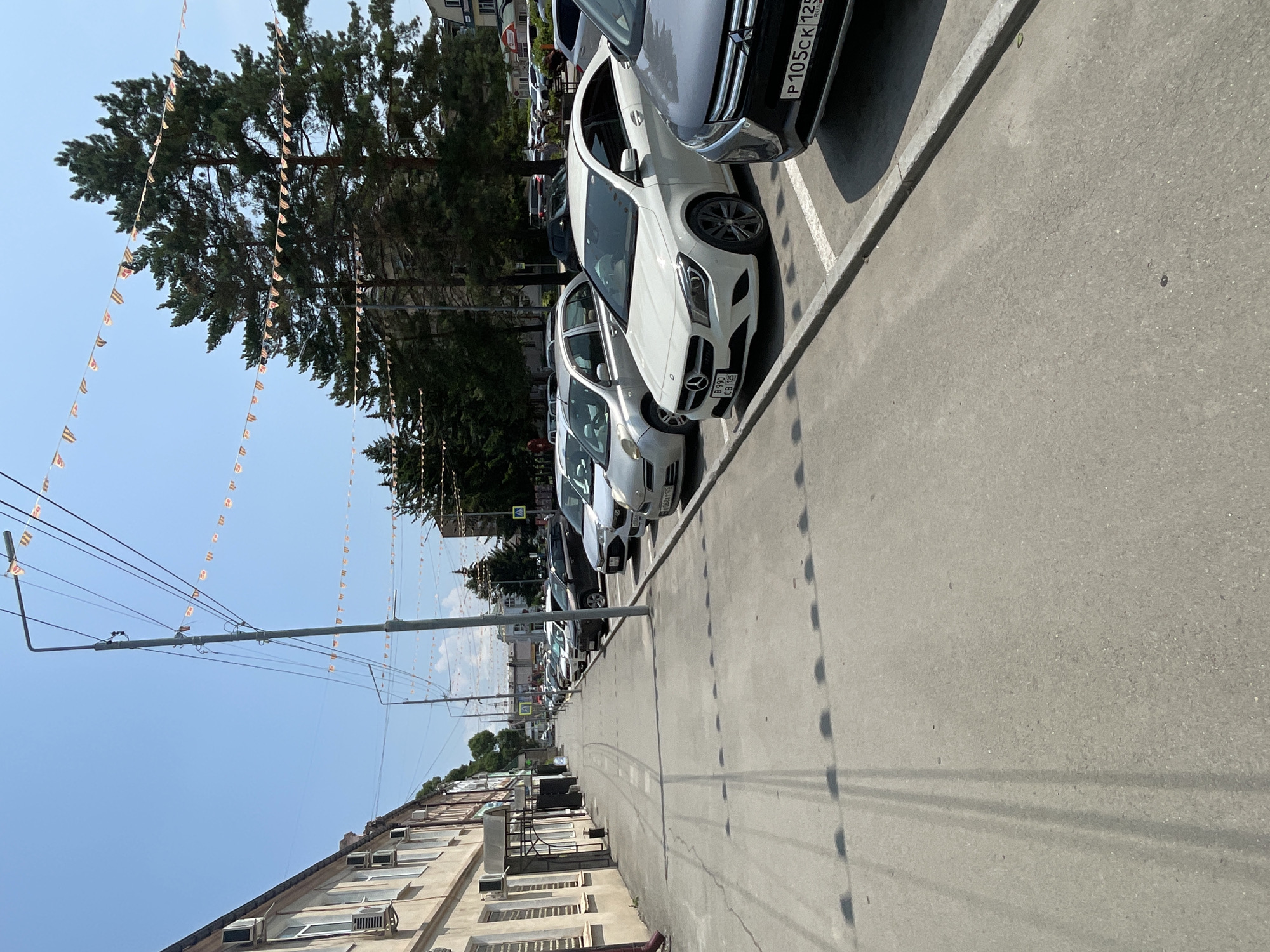 The image depicts a street scene in Spassk-Dal'nij, Russia. The perspective is from a low angle, looking up towards a clear blue sky and across a row of parked cars. The cars are lined up on a paved street, with white parking lines visible. In the foreground, the side of a building with multiple windows and air conditioning units can be seen. Overhead, wires are strung across the scene, and a line of small, colorful triangular flags is suspended between poles, suggesting a festive occasion or decoration.
The subjects in the image are primarily cars, with the suggestion of people inside some of them. The setting is an urban environment, likely a street or parking area in a town. The activity taking place is simply the presence of parked vehicles on a street on a sunny day.
Notable details include the bright sunlight, indicating it is daytime, likely mid-morning or afternoon. The clear sky suggests fair weather. The building in the foreground provides some architectural context, while the flags add a touch of color and occasion. Some text is visible on the license plates of the cars: one plate reads "P 105CK 125 RUS" and another appears to be "B 090 CB 125". The overall impression is a calm, ordinary street scene with hints of a celebration.