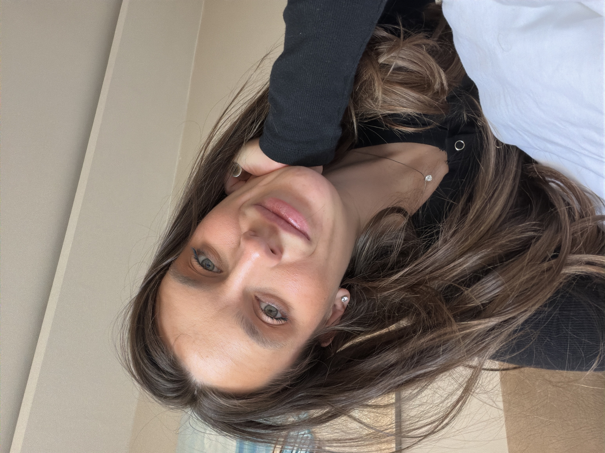 A woman with long brown hair and fair skin is pictured from a slightly downward, rotated angle. She is wearing a black top and a delicate necklace with a single diamond pendant. Her left hand is near her face, touching her cheek. She appears to have light eye makeup and is wearing a small earring. A corner of what seems to be a white pillow is visible in the upper right corner. The background features a plain, light-colored wall. The location in the city of Budva, Montenegro cannot be determined from this image.