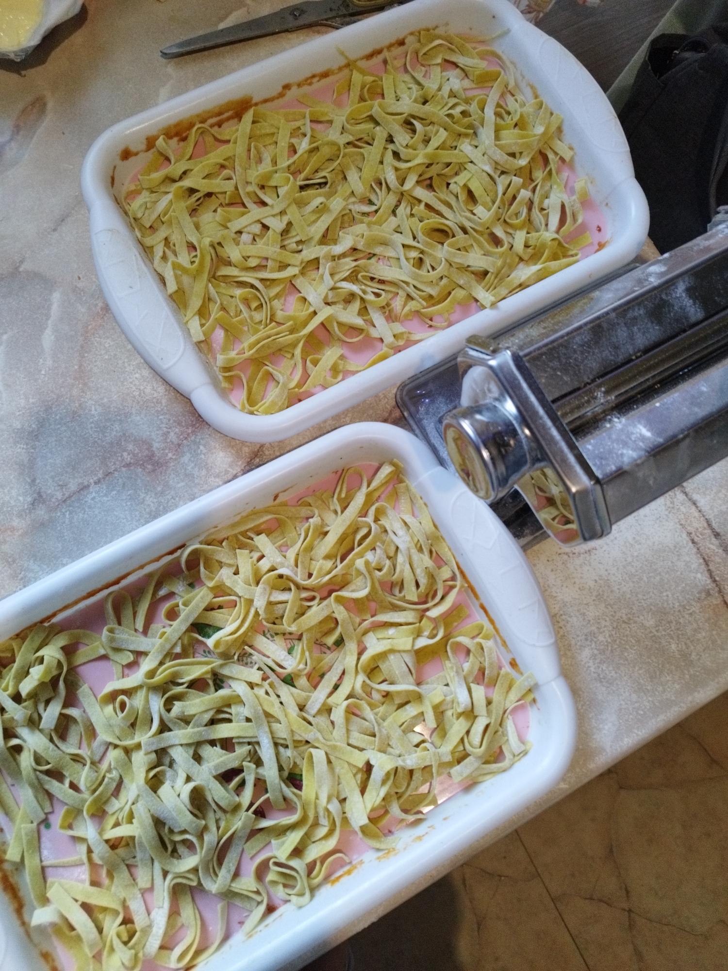 Two white rectangular trays are filled with freshly made pasta. The pasta appears to be tagliatelle or a similar flat noodle, a light yellow-green in color, and is scattered unevenly within the trays. A manual pasta machine, made of silver metal, is positioned on the right side of the image, partially obscuring the top tray. The machine has a crank and rollers visible. The surface beneath the trays appears to be a countertop with a marble-like pattern in shades of beige and grey. In the background, a pair of scissors with grey handles lies on the countertop near the top edge of the image. There is no indication of the city of Lozova, Ukraine, in the image.